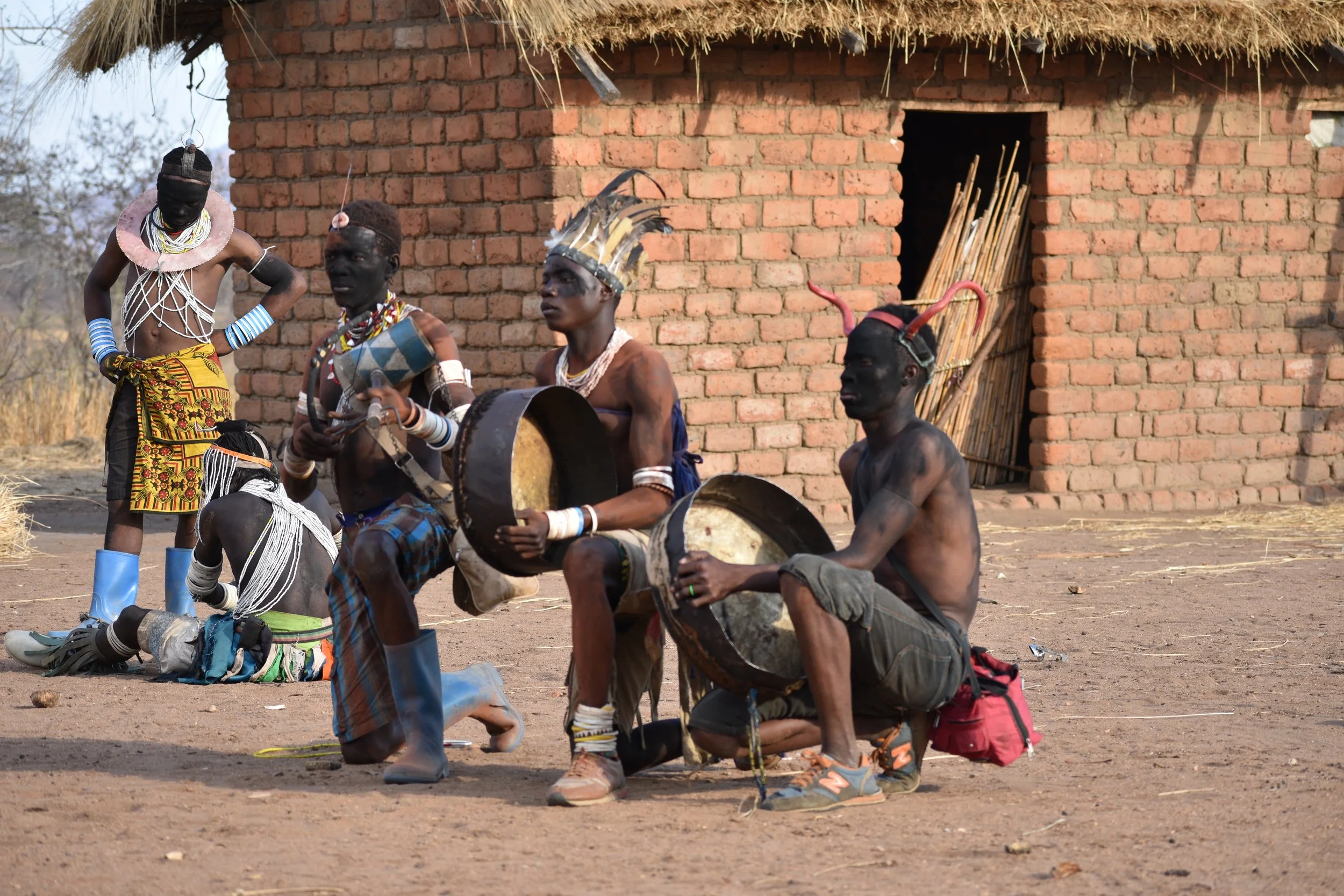 A group of indigenous people, likely from Africa, dressed in traditional attire, engaged in a ritual or dance outside a brick building with a straw roof. Some are seated, holding drums, while others are standing, wearing elaborate jewelry and headdre