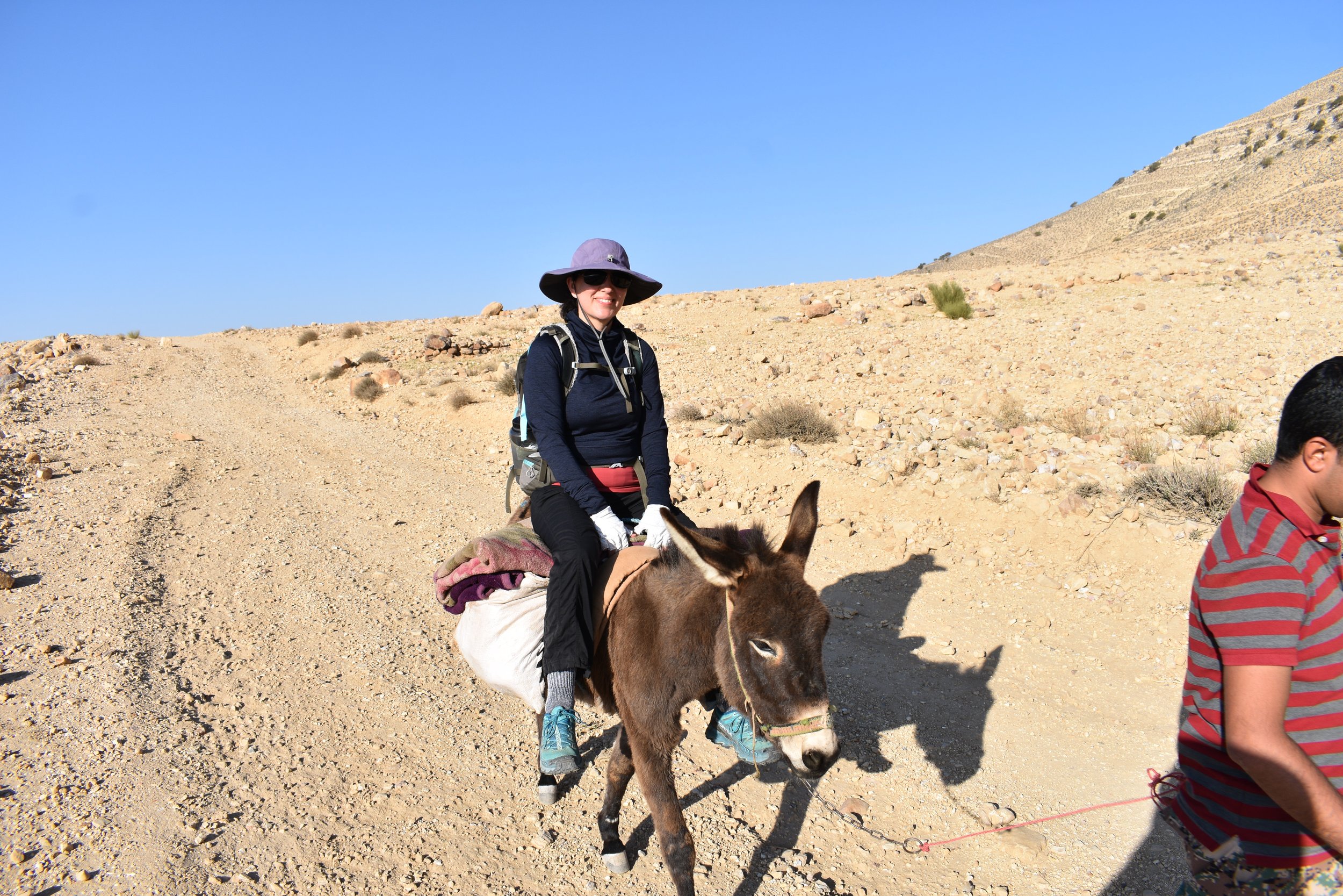 A person wearing a sun hat rides a donkey on a dirt path in a desert landscape, accompanied by a person in a striped shirt walking next to them.