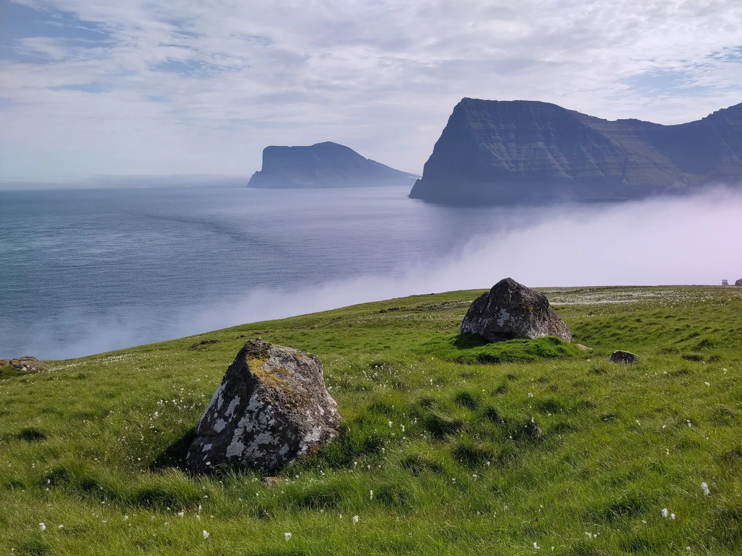 Green grassy hillside with large rocks, overlooking an ocean with cliffs in the distance, mist or fog near the water, under a partly cloudy sky.