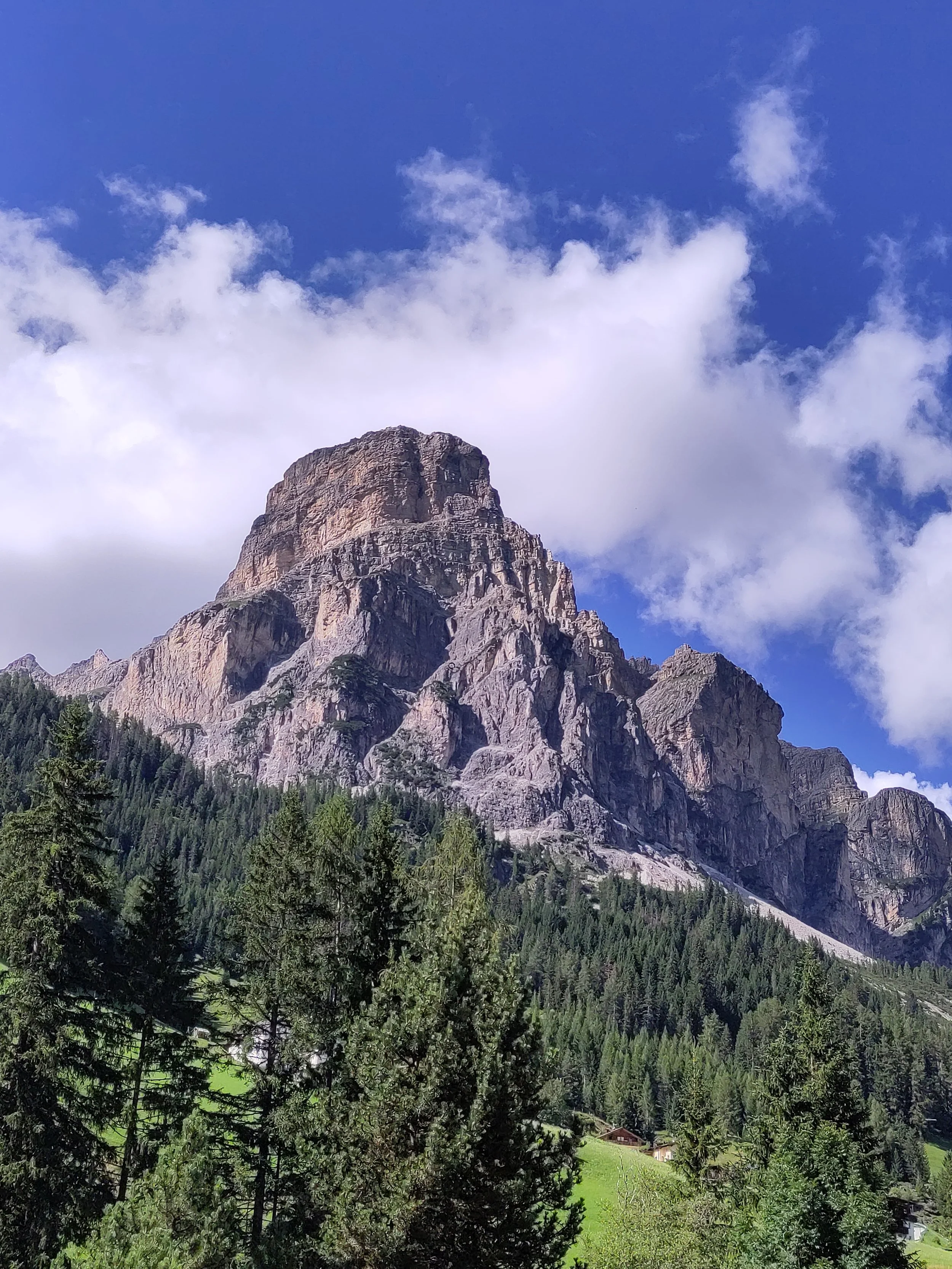 A tall mountain with rocky cliffs and a forested lower slope under a partly cloudy blue sky.