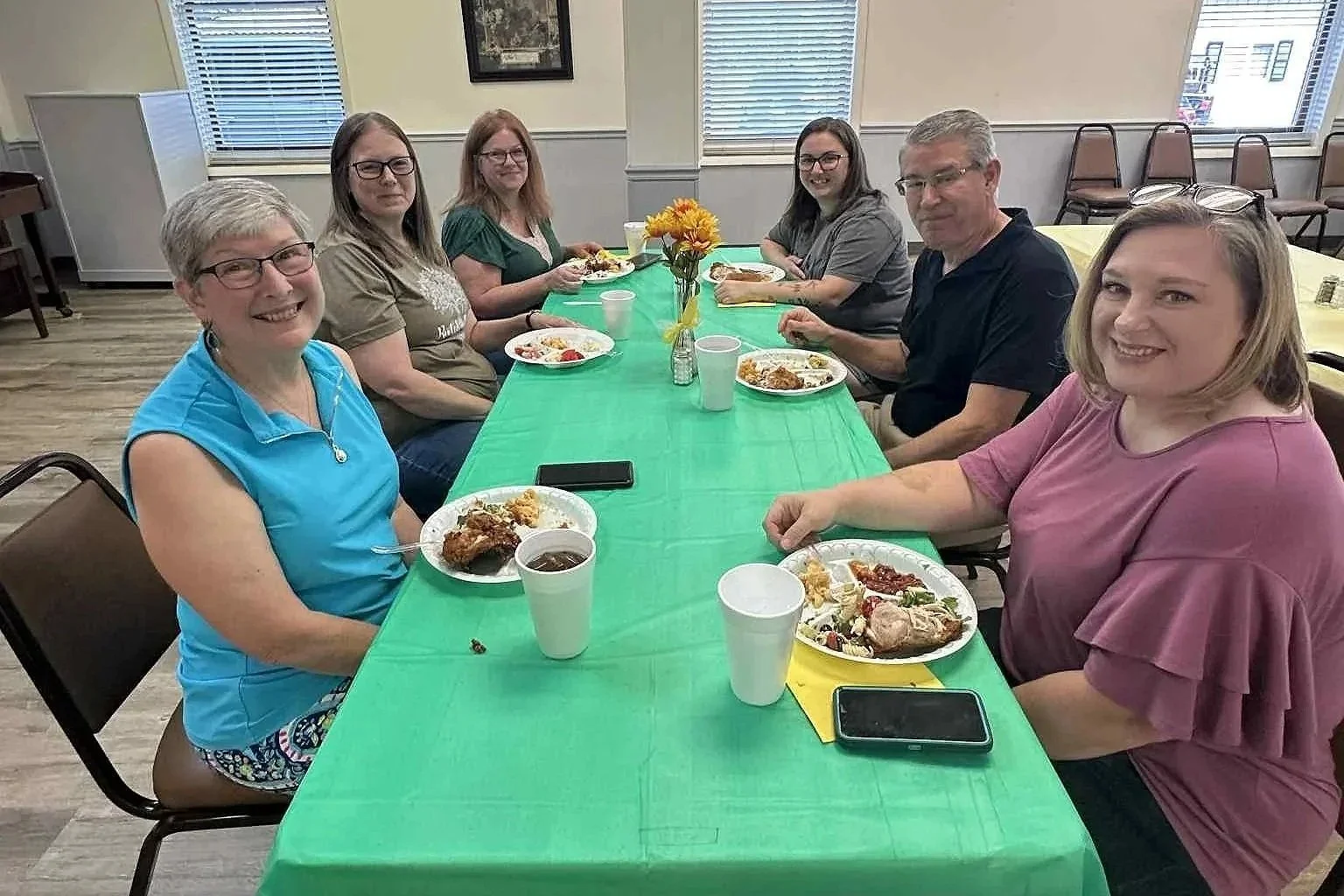 People sitting at a long table with green tablecloth, having a meal in a bright room with windows, a small flower bouquet in the center of the table.