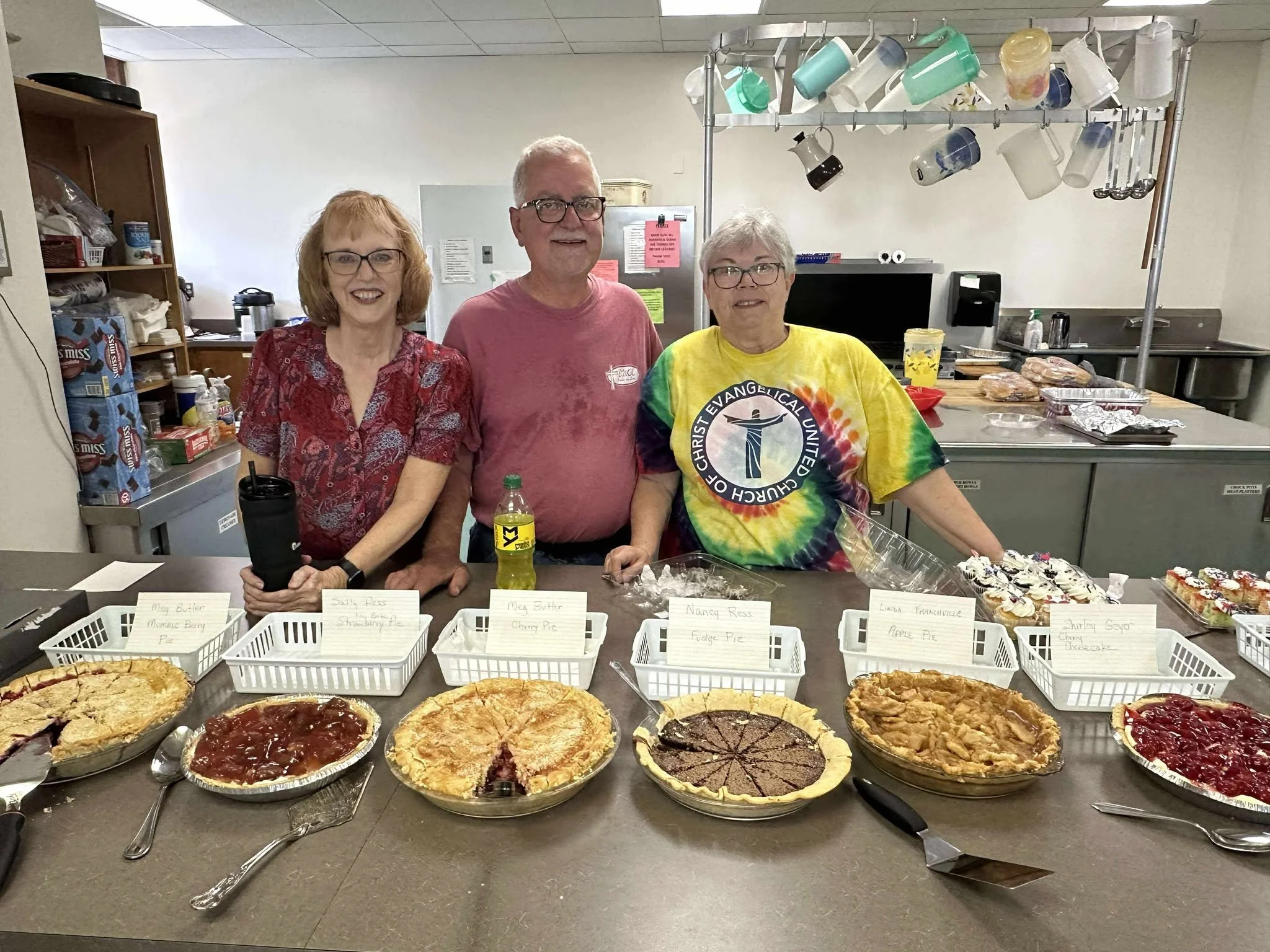 Three smiling women and a man standing behind a dessert table with various homemade pies. The women are holding a black insulated tumbler, a yellow soda bottle, and a clear plastic bag. The table displays five different pies, including cherry, apple,