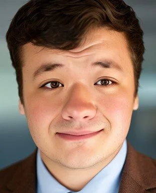 Close-up of a young man with brown hair, one eye winking, and a slight smile, wearing a blue shirt and brown blazer.
