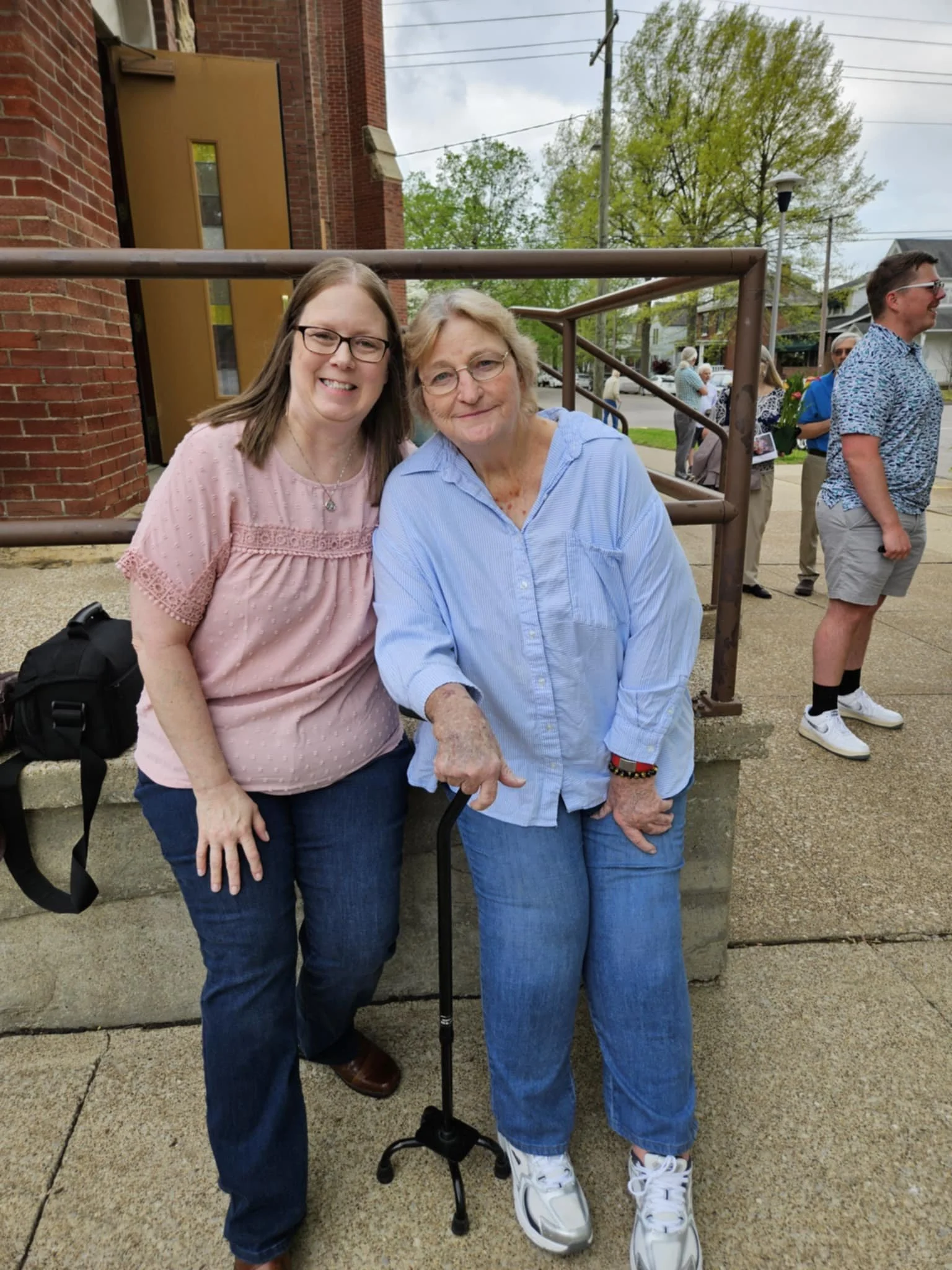 Two women standing outdoors, smiling for the camera; one young woman with glasses, and one woman with a cane, wearing casual clothes and sneakers.