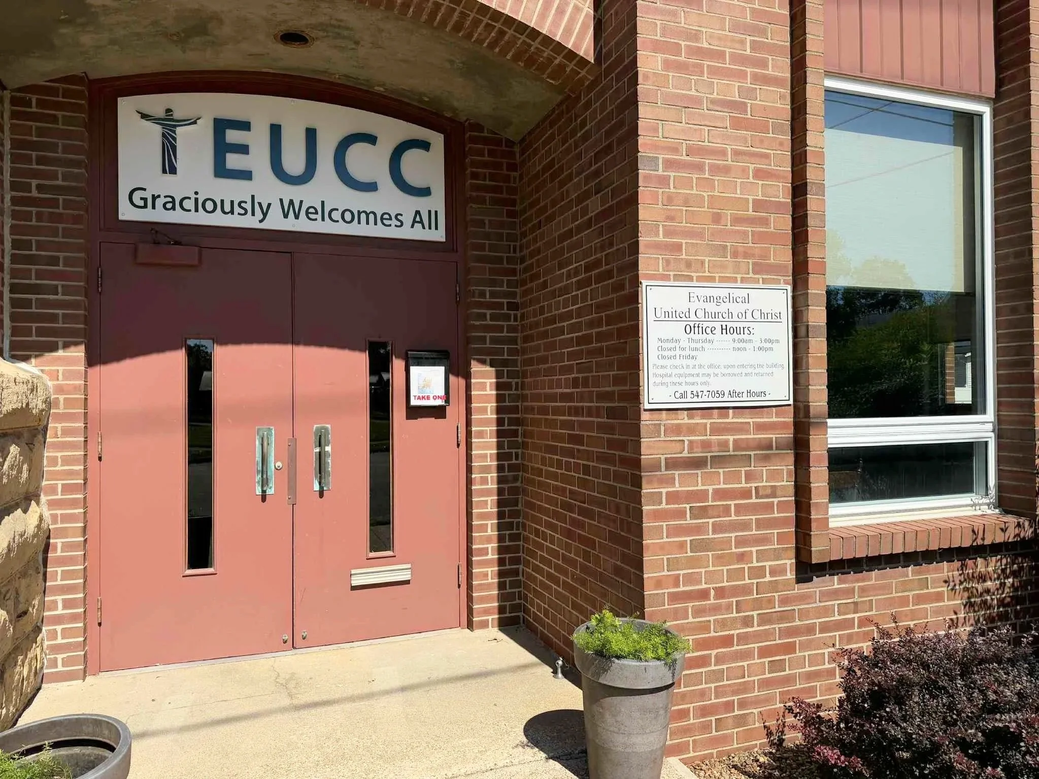 Entrance of a brick church with a sign that reads 'TEUC Graciously Welcomes All.' A side sign displays office hours for the Evangelical United Church of Christ, featuring days and times open and contact information.