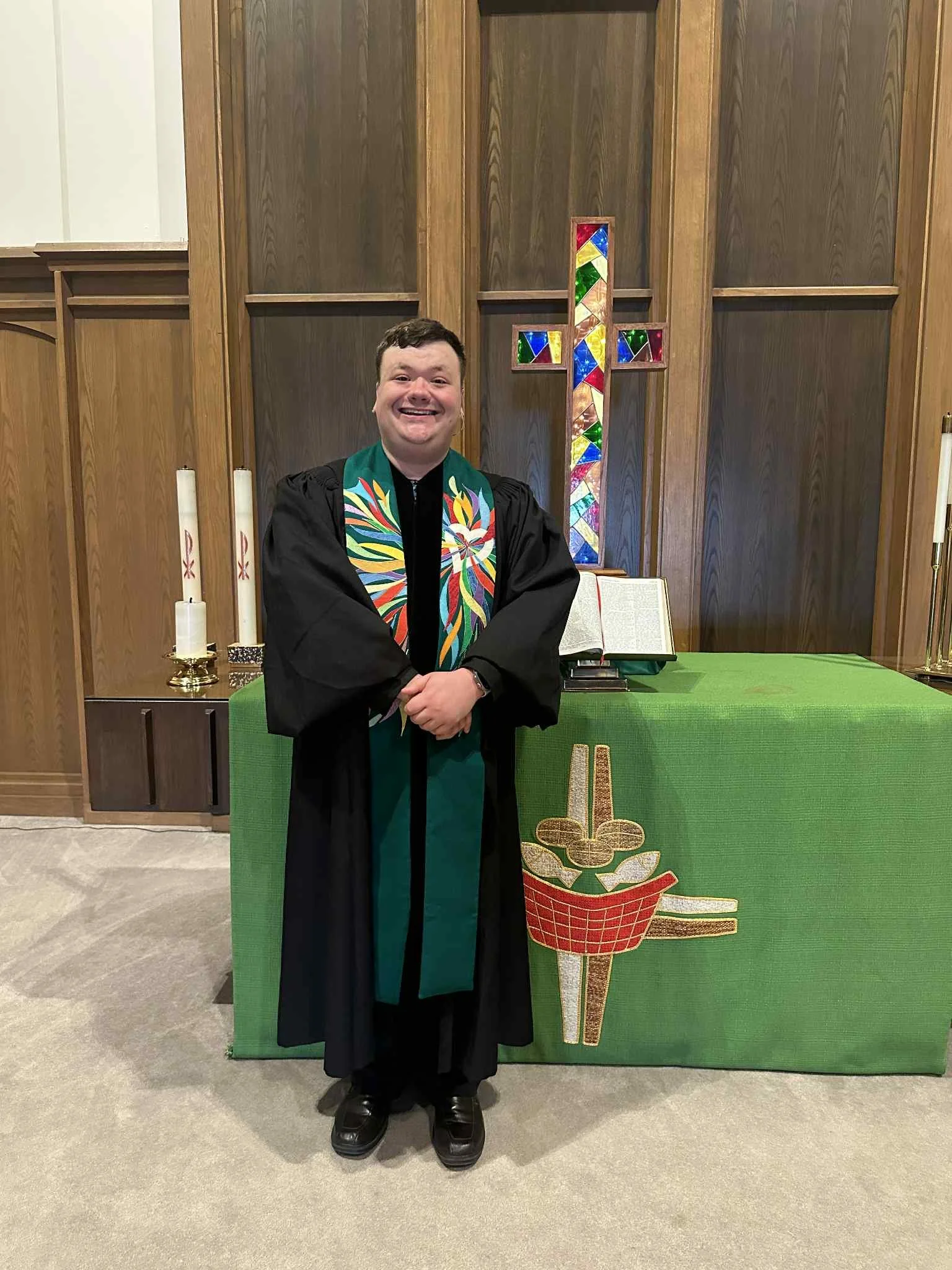 A man in a black clergy robe and colorful stole smiling and standing in front of a church altar with a stained-glass cross, candles, and an open Bible.