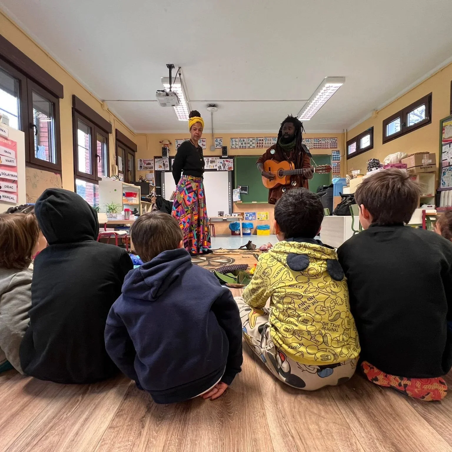 Niños sentados en el suelo de un aula viendo una presentación artística, con dos personas en el escenario tocando guitarra y cantando, una de ellas vestida con ropa colorida y la otra con ropa oscura, en un aula escolar decorada con posters y ventana