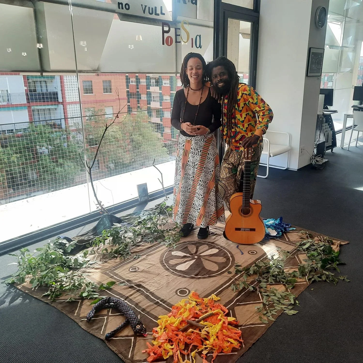 Dos personas con vestimenta colorida y sonriendo, una de ellas sostiene una guitarra. Están en un espacio interior con mucha luz natural, decorado con plantas y objetos como un tapiz, ramas, y fuego artificial de colores en el suelo, creando un ambie
