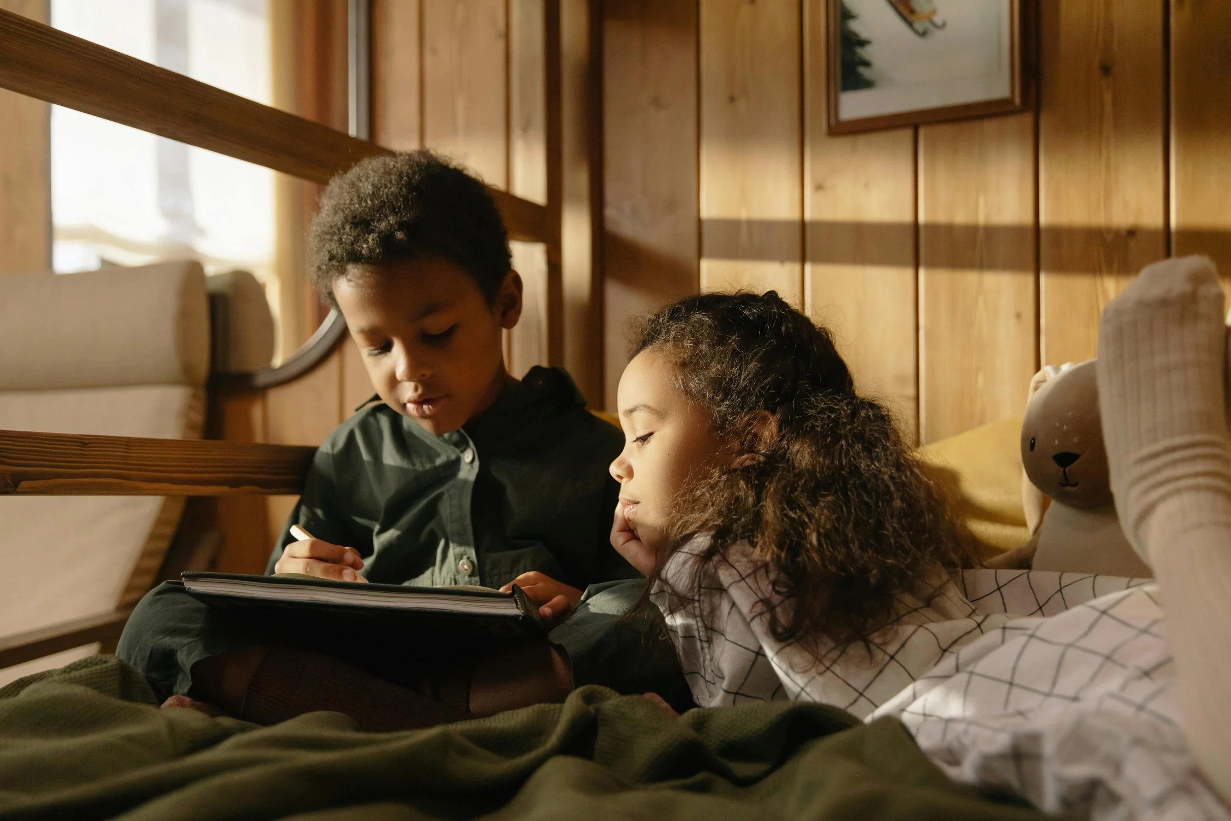 Dos niños, uno rubio y otro oscuro, leyendo un libro juntos en una cama en una habitación de madera con luz natural