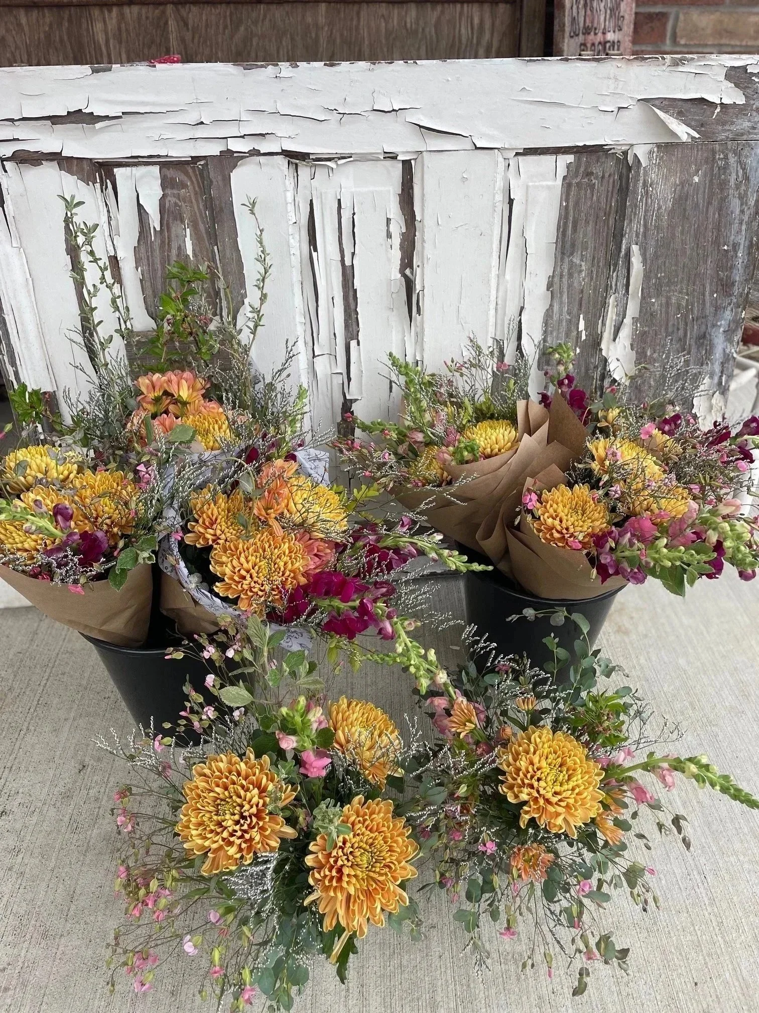 Four bouquets of different flowers in black and brown wrapping paper, placed on a table in front of a distressed white wooden backdrop.
