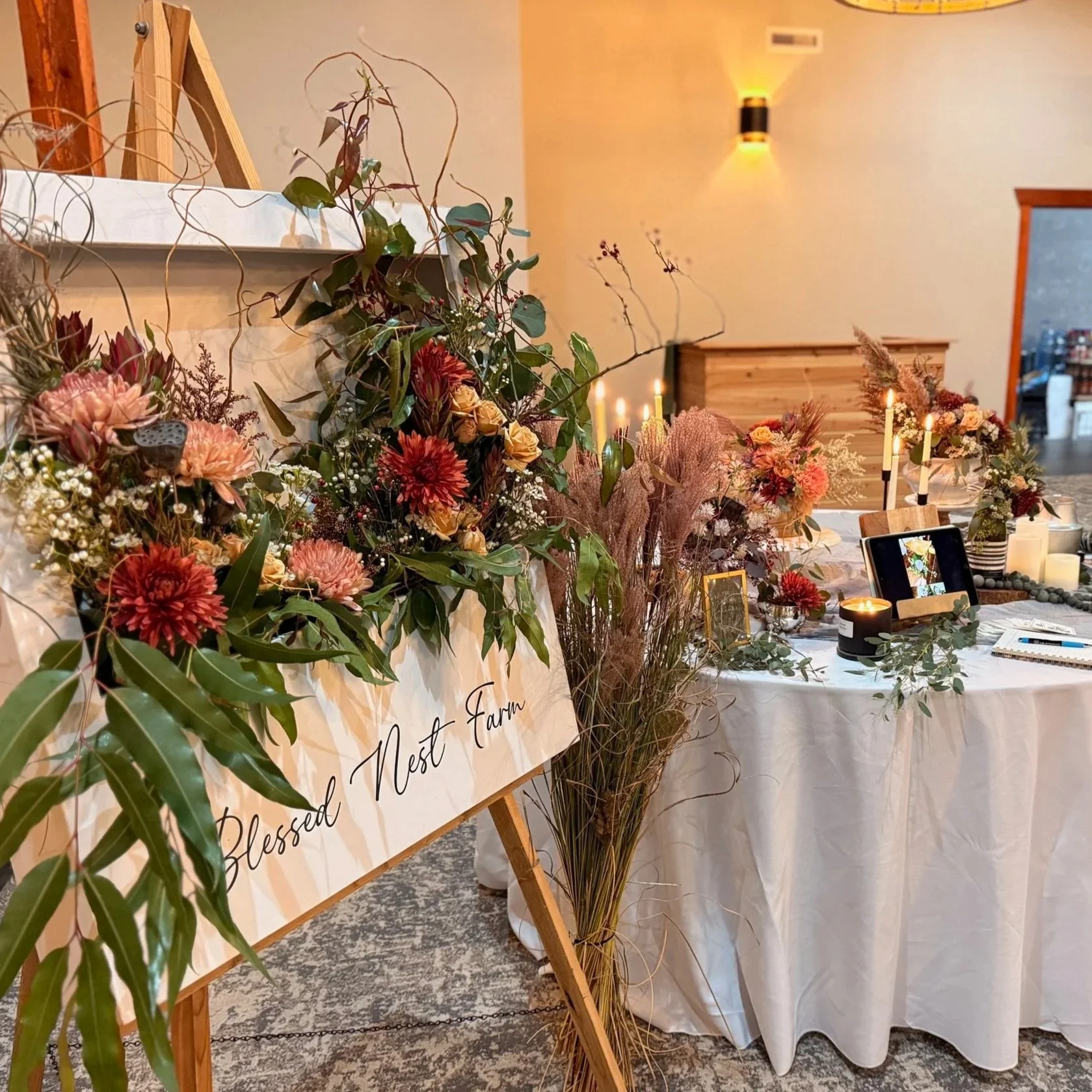 Decorative floral arrangements with pink, red, and peach flowers, greenery, and candles on a wedding or event table with a sign reading 'Blessed Nest Farm' in a warmly lit indoor setting.