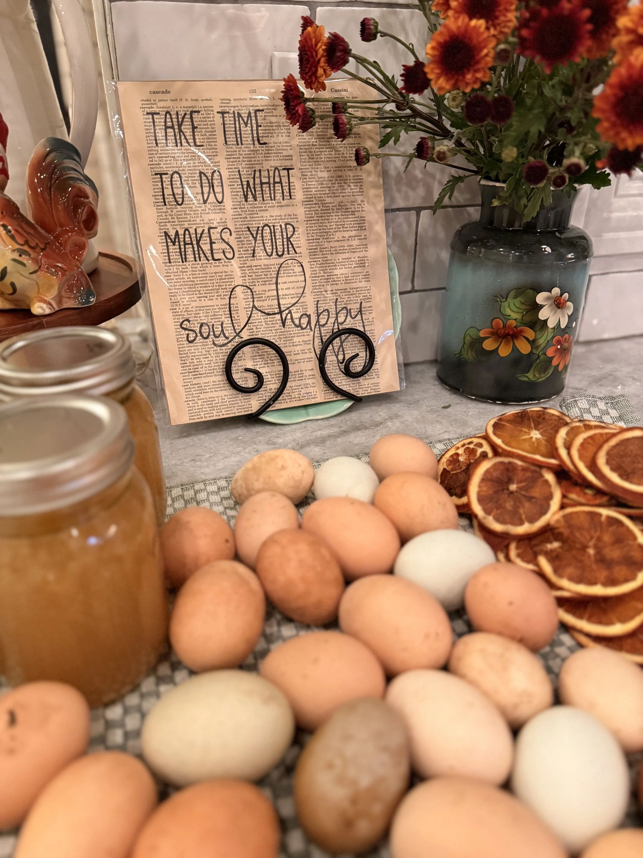 A decorative display with a variety of eggs, dried orange slices, and a jar of honey on a table. Behind the display, there is a sign that says, 'Make time to do what makes your soul happy,' with a background of printed text and a vase with orange and white flowers.