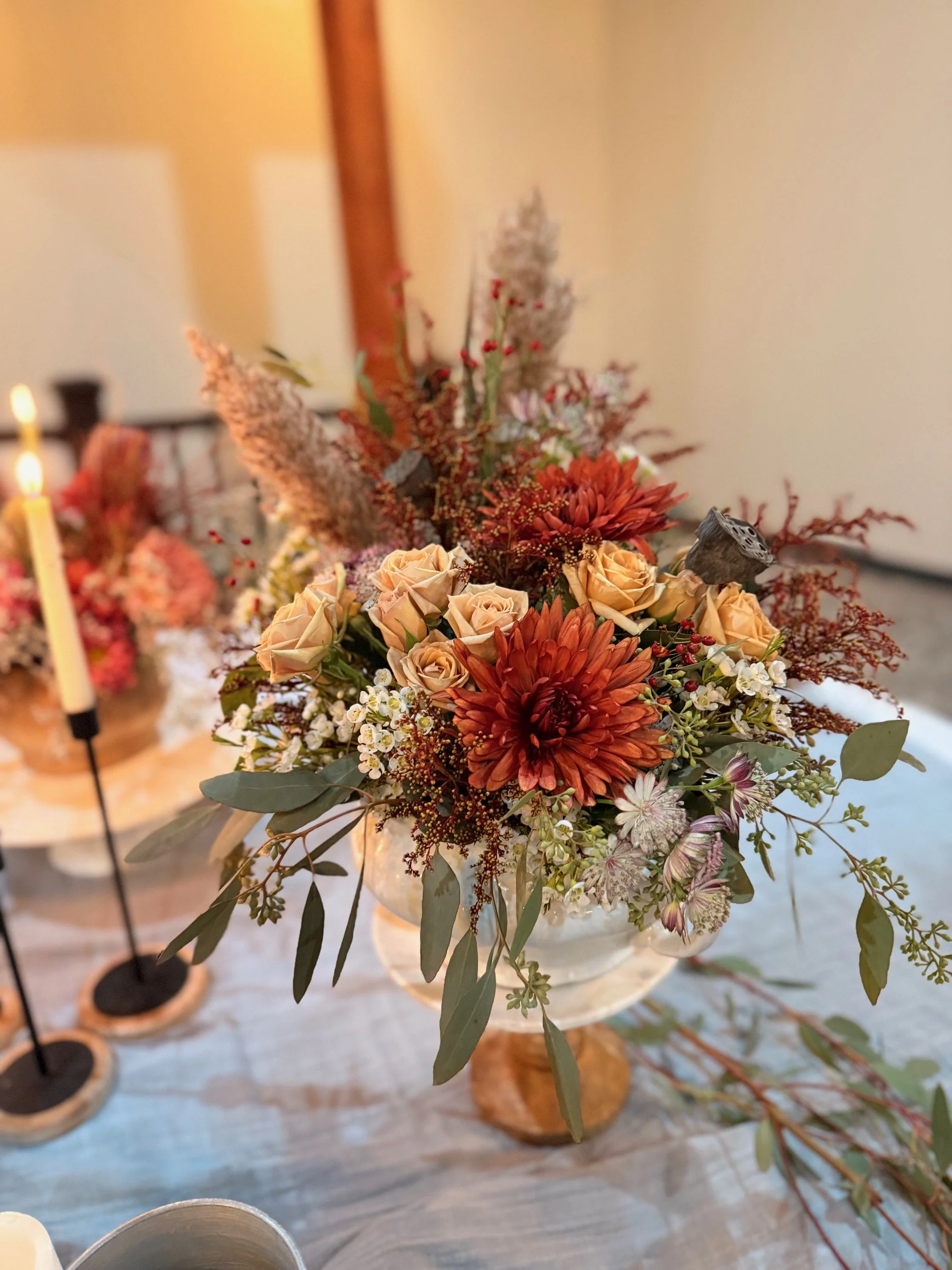 A floral centerpiece with orange and peach roses, rust-colored dahlias, white and pink flowers, and greenery in a glass vase on a table with additional flower arrangements and candles.