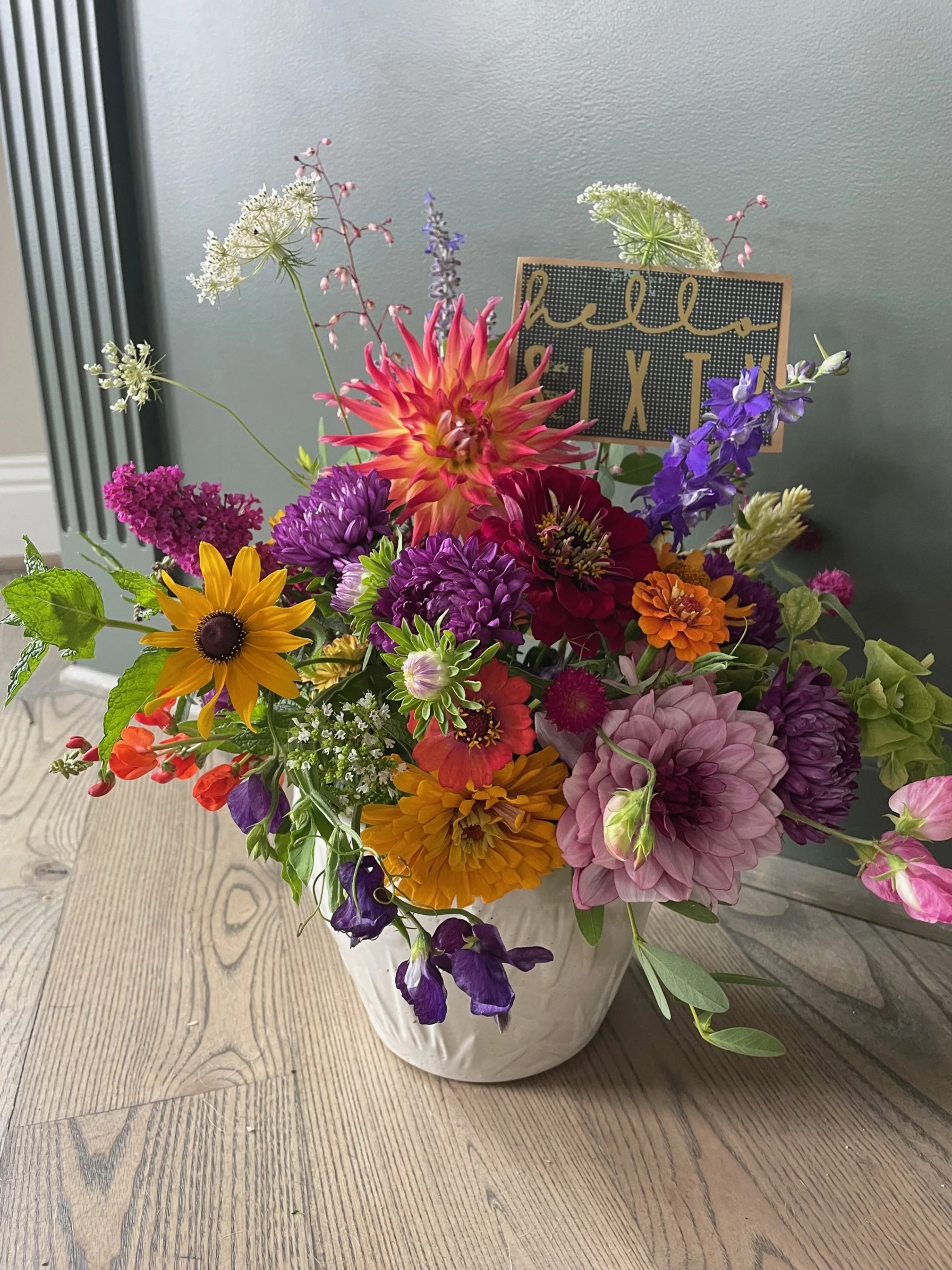 A colorful bouquet of various flowers in a white vase on a wooden table, with a small sign that reads 'hello relax' in the background.