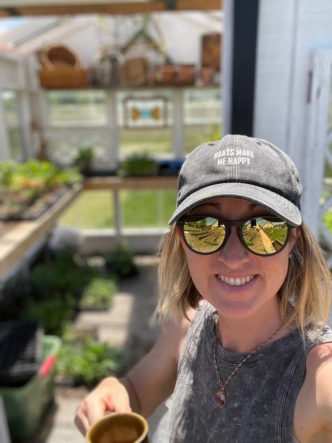 A woman wearing a gray baseball cap with the words 'Goats Make Me Happy', reflective sunglasses, and a necklace, smiling in a greenhouse with plants and gardening tools around her.