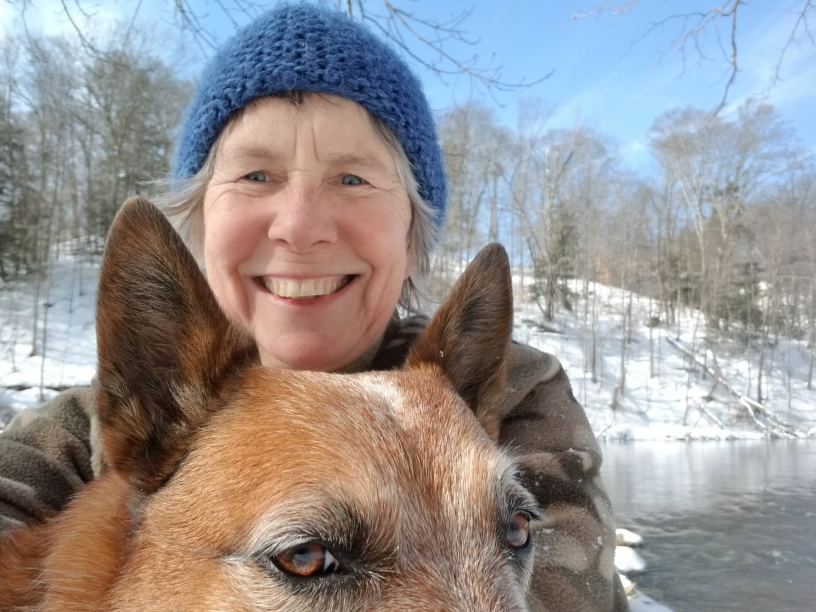 Jana wearing a blue knit hat and a brown jacket smiles while holding a dog. The background features a snowy landscape with trees and a partially frozen river under a clear blue sky.