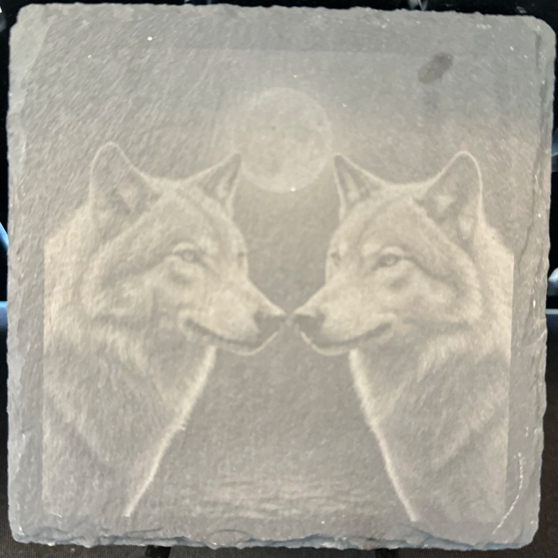 A engraved slate coaster featuring two wolves facing each other with a moon above their heads.