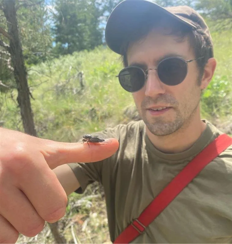 A man wearing sunglasses, a cap, and a gray t-shirt points his finger at a small insect on his finger in a forested area.