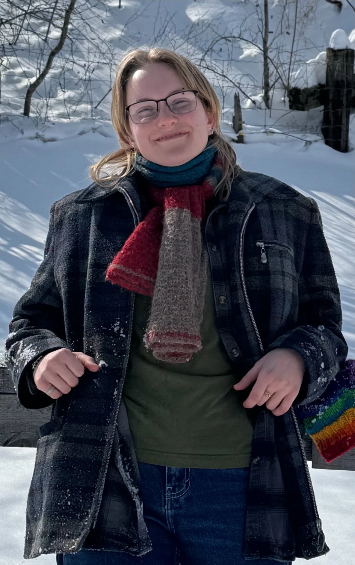 A woman with glasses and shoulder-length hair stands outdoors in snowy weather, smiling, wearing a black jacket, a green shirt, a scarf, and colorful gloves.