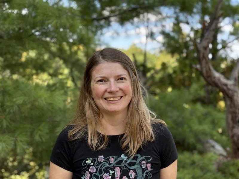A smiling woman with long light brown hair standing outdoors with green trees and foliage in the background.