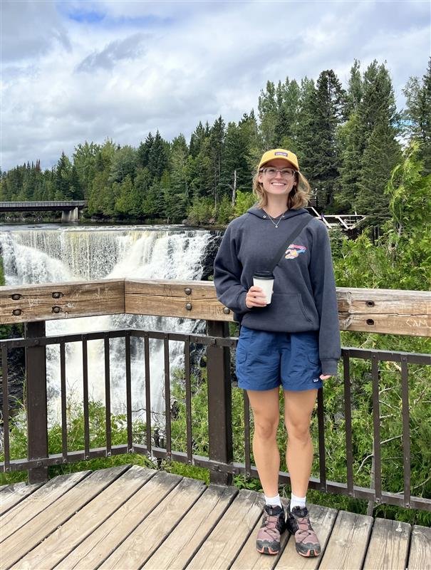 A young woman standing on a wooden observation deck, holding a paper cup, with a waterfall and dense forest in the background on a cloudy day.