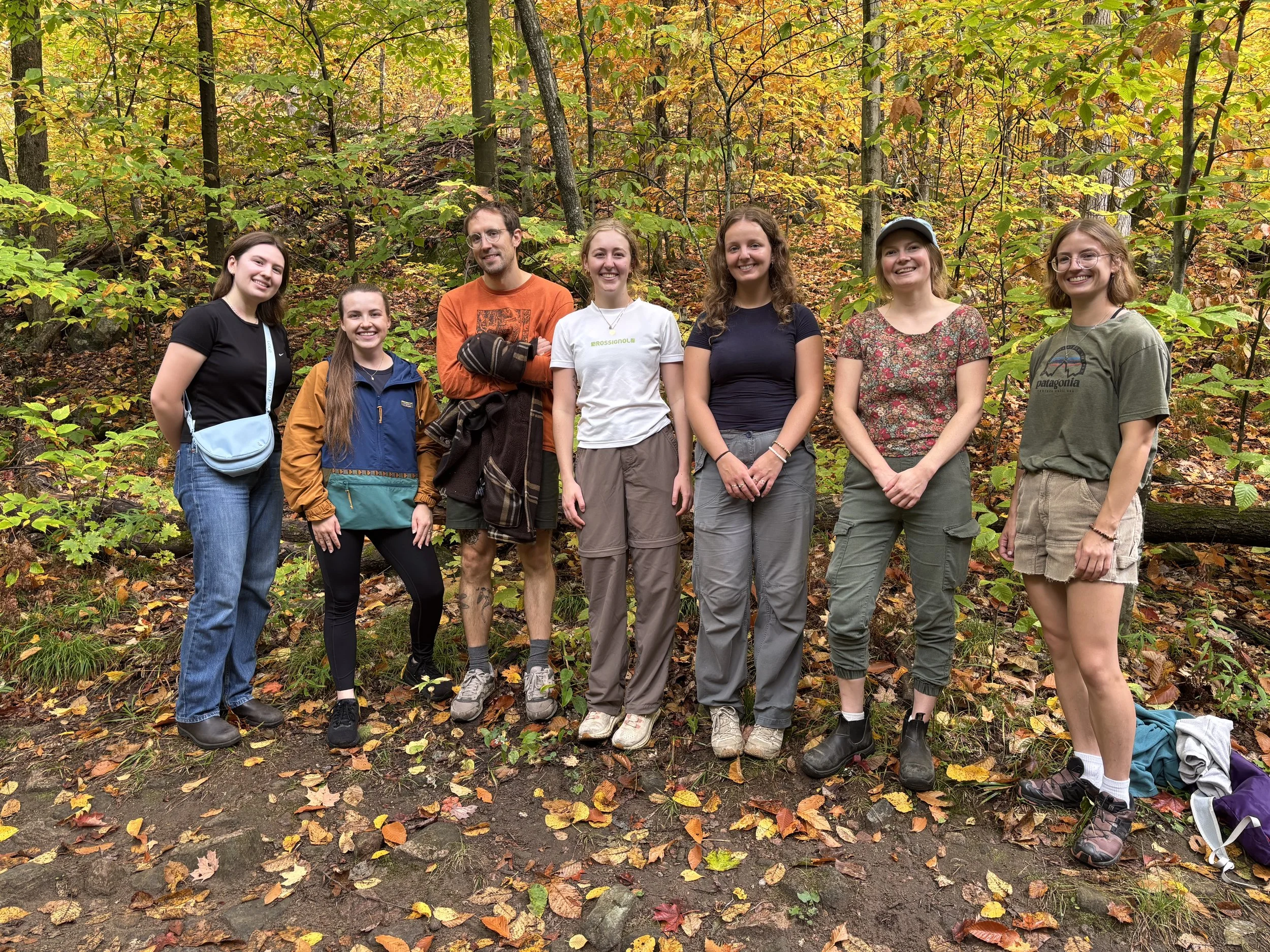 Group of seven young adults standing on a forest path surrounded by autumn-colored trees, smiling and relaxing during a hike.