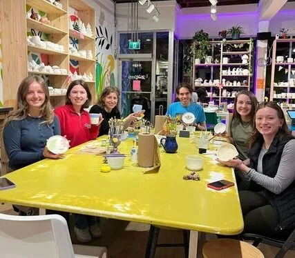 Six women sitting around a yellow table in a pottery painting studio, holding painted pottery pieces and smiling.