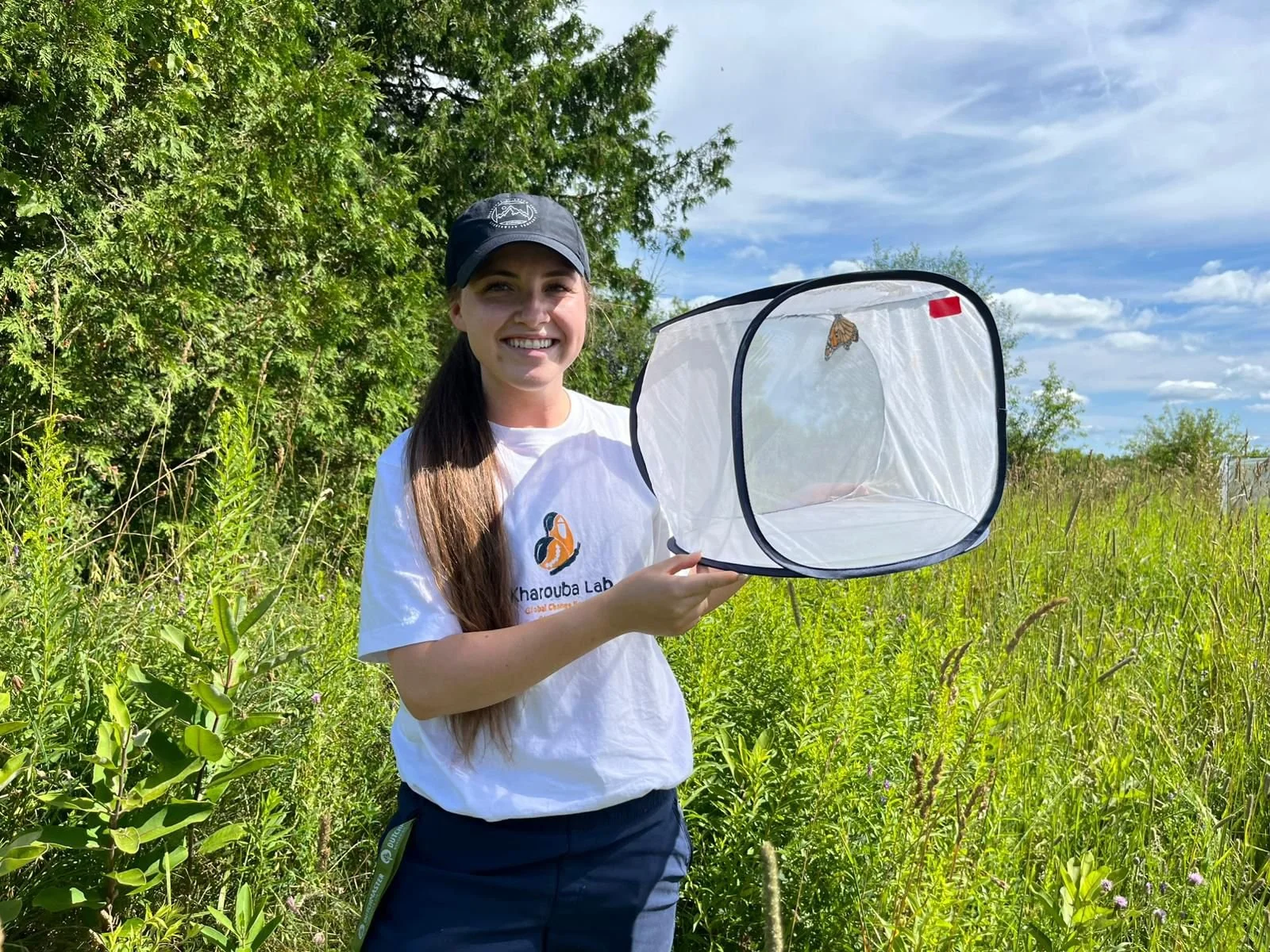 A woman smiling outdoors holding a butterfly net in a grassy field with trees and a blue sky in the background.
