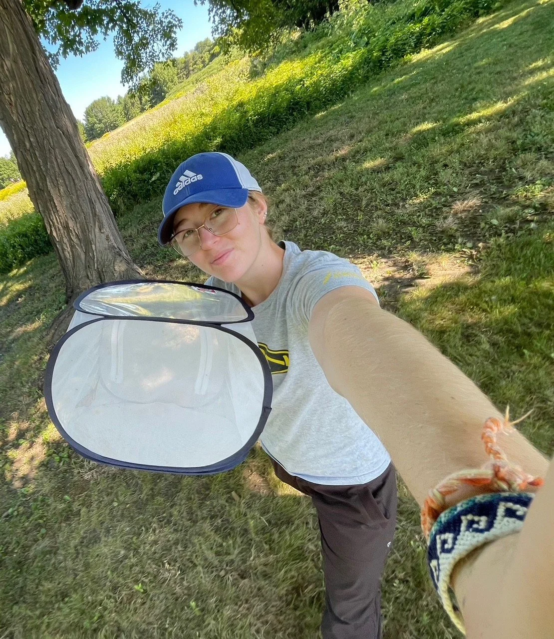 A young woman with glasses, wearing a blue Adidas cap and a gray t-shirt with yellow accents, taking a selfie outdoors under a tree. She is holding an open clear plastic display case, standing on a grassy area with trees and shrubs in the background on a sunny day.