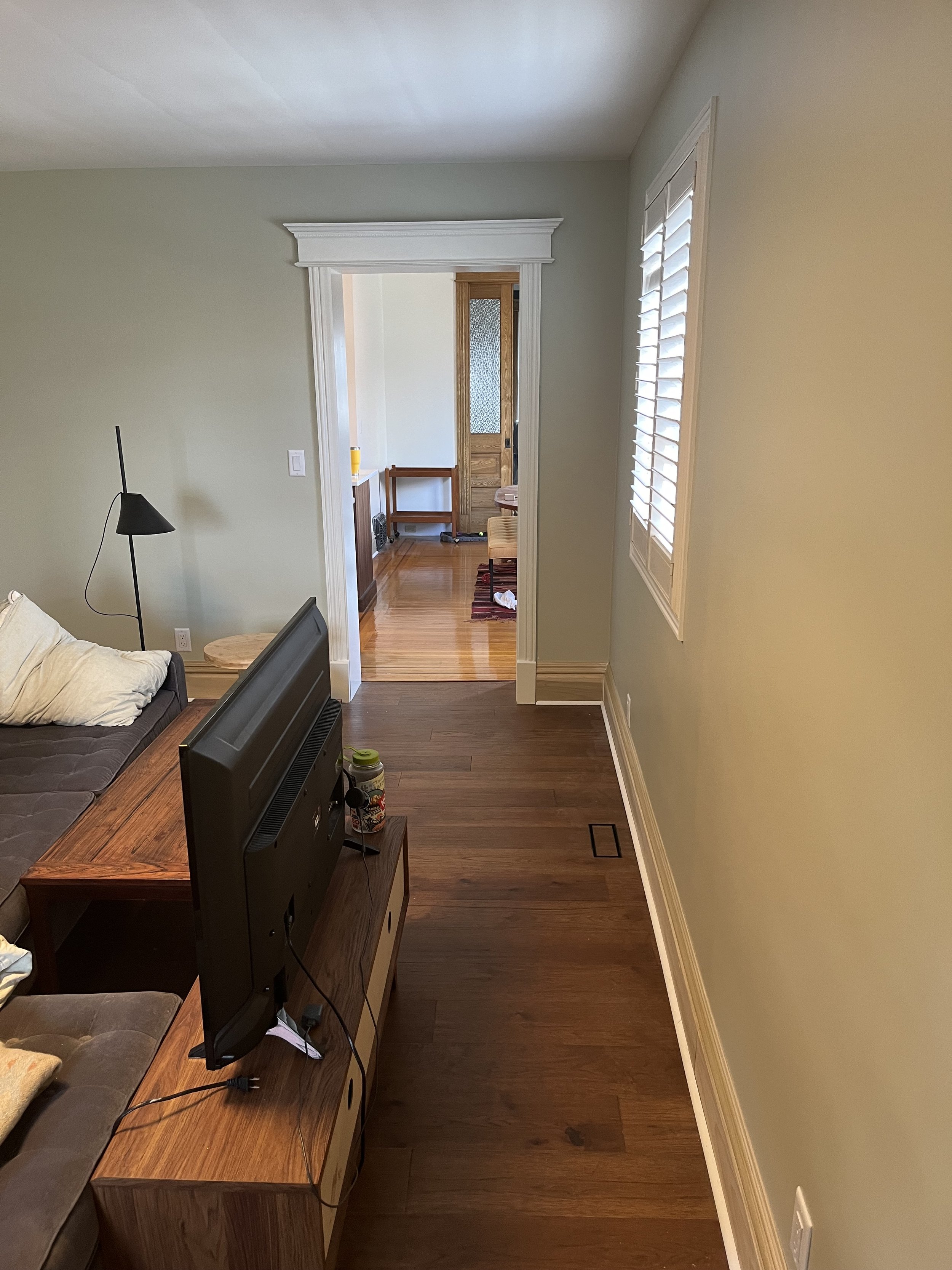 Interior view of a living room with a TV on a stand, a couch with a pillow, and a doorway leading to another room. The floor has dark wooden planks and there's a window with white shutters.