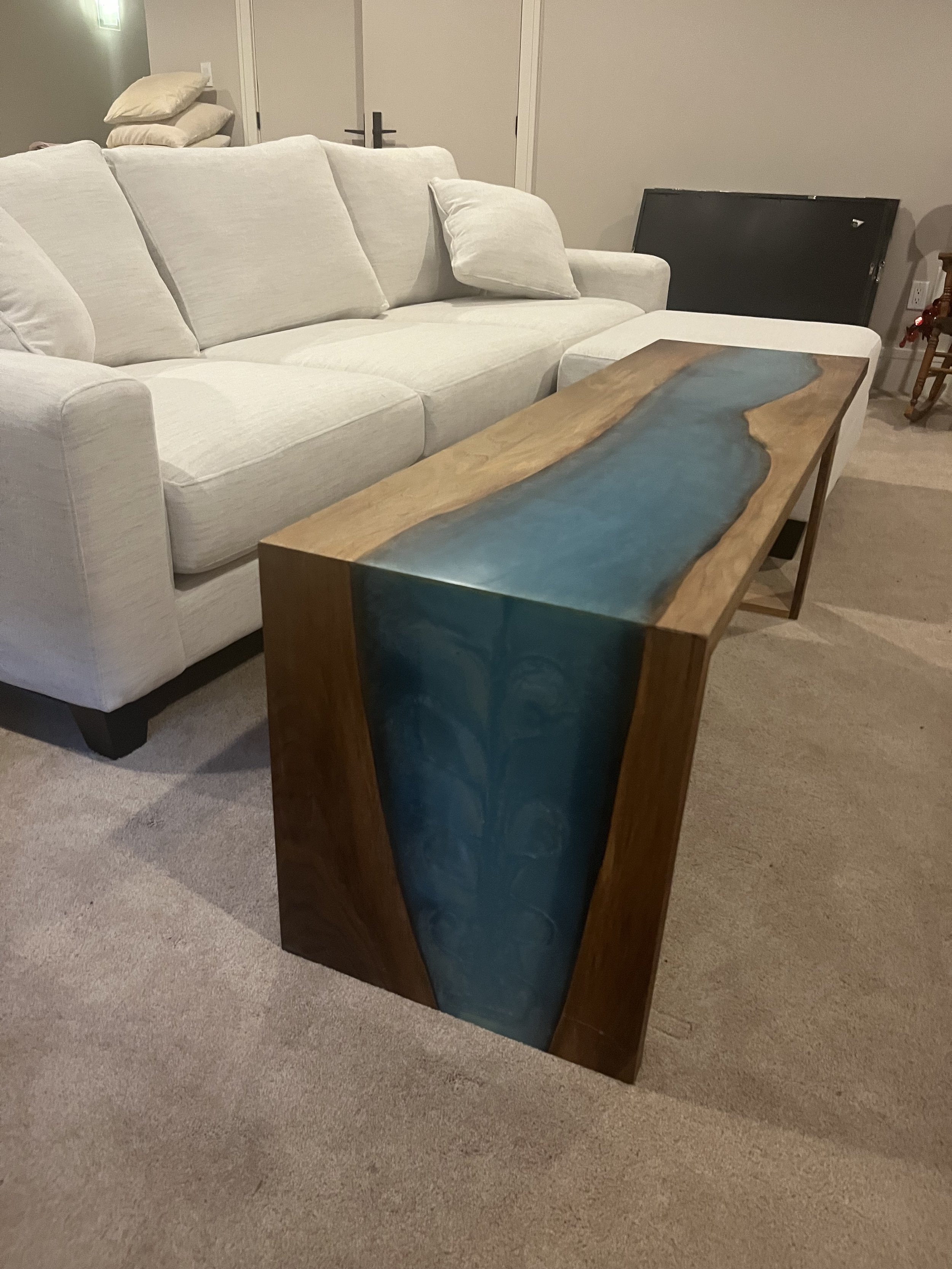 Interior room with a light gray sofa and a wooden coffee table featuring a blue resin river design on a beige carpet.