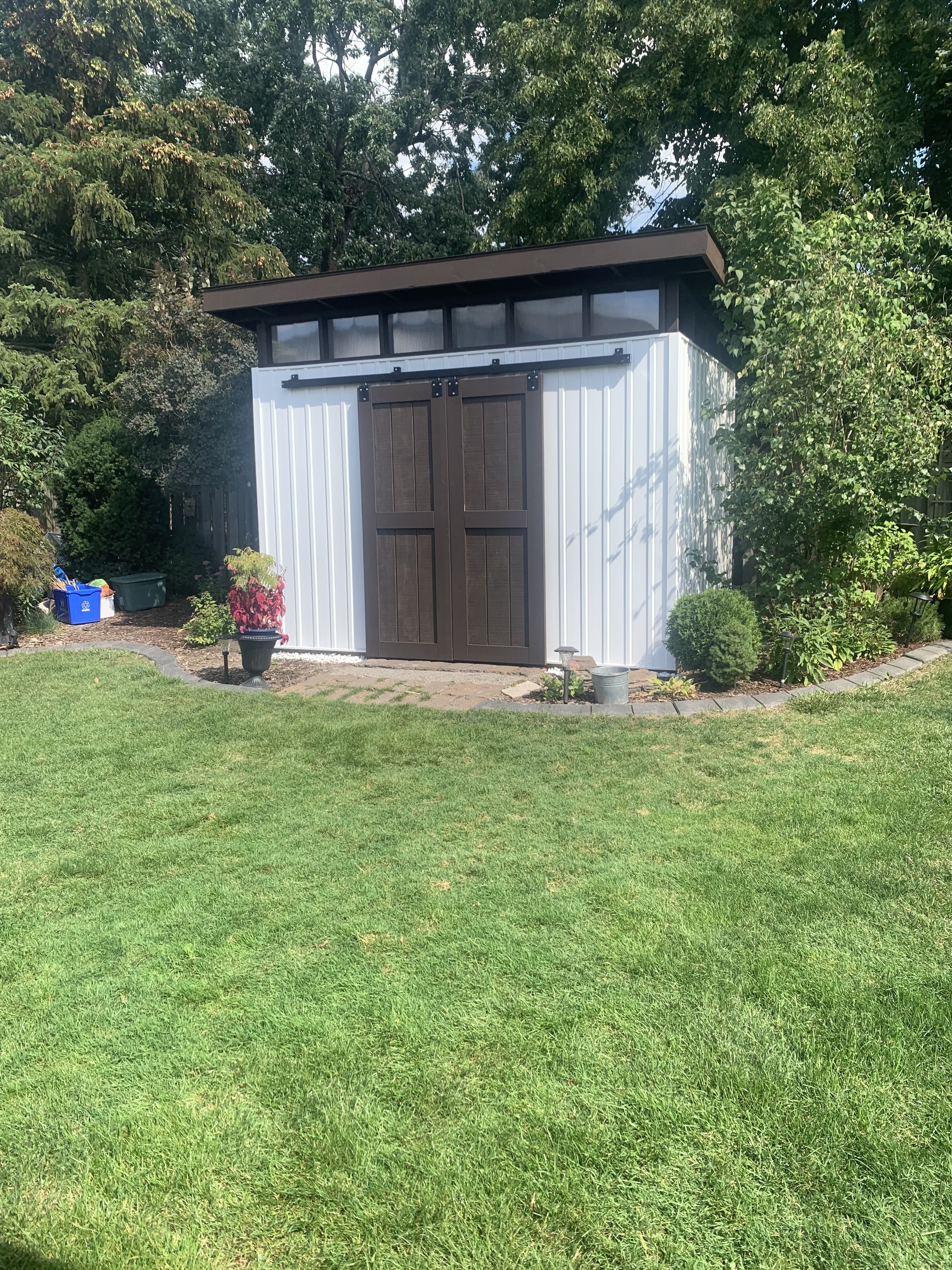 Backyard shed with white siding and dark brown double doors, surrounded by greenery and grass lawn on a sunny day.