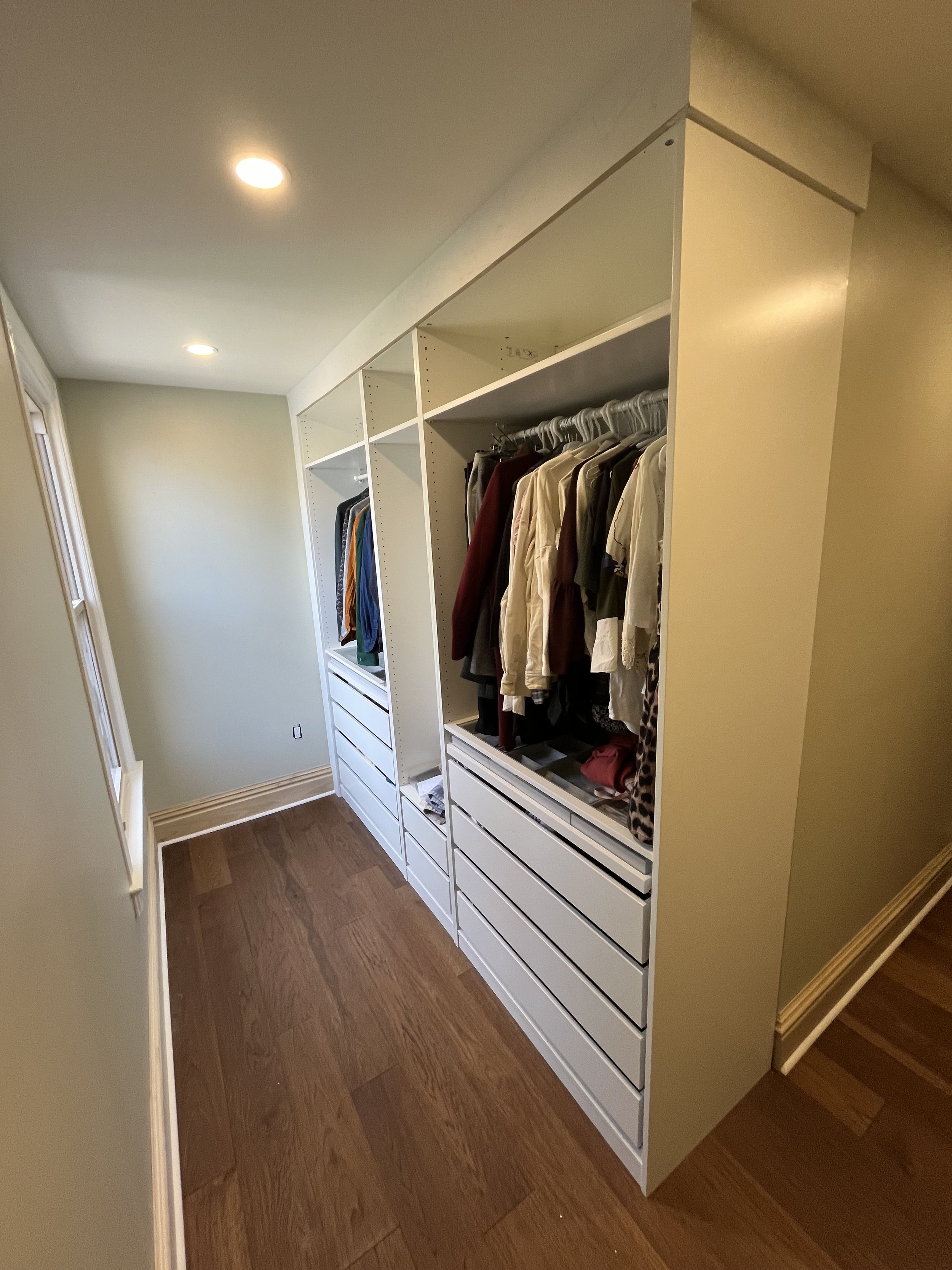 A neatly organized walk-in closet with white shelving units, hanging clothes, and multiple drawers, placed on a wooden floor.