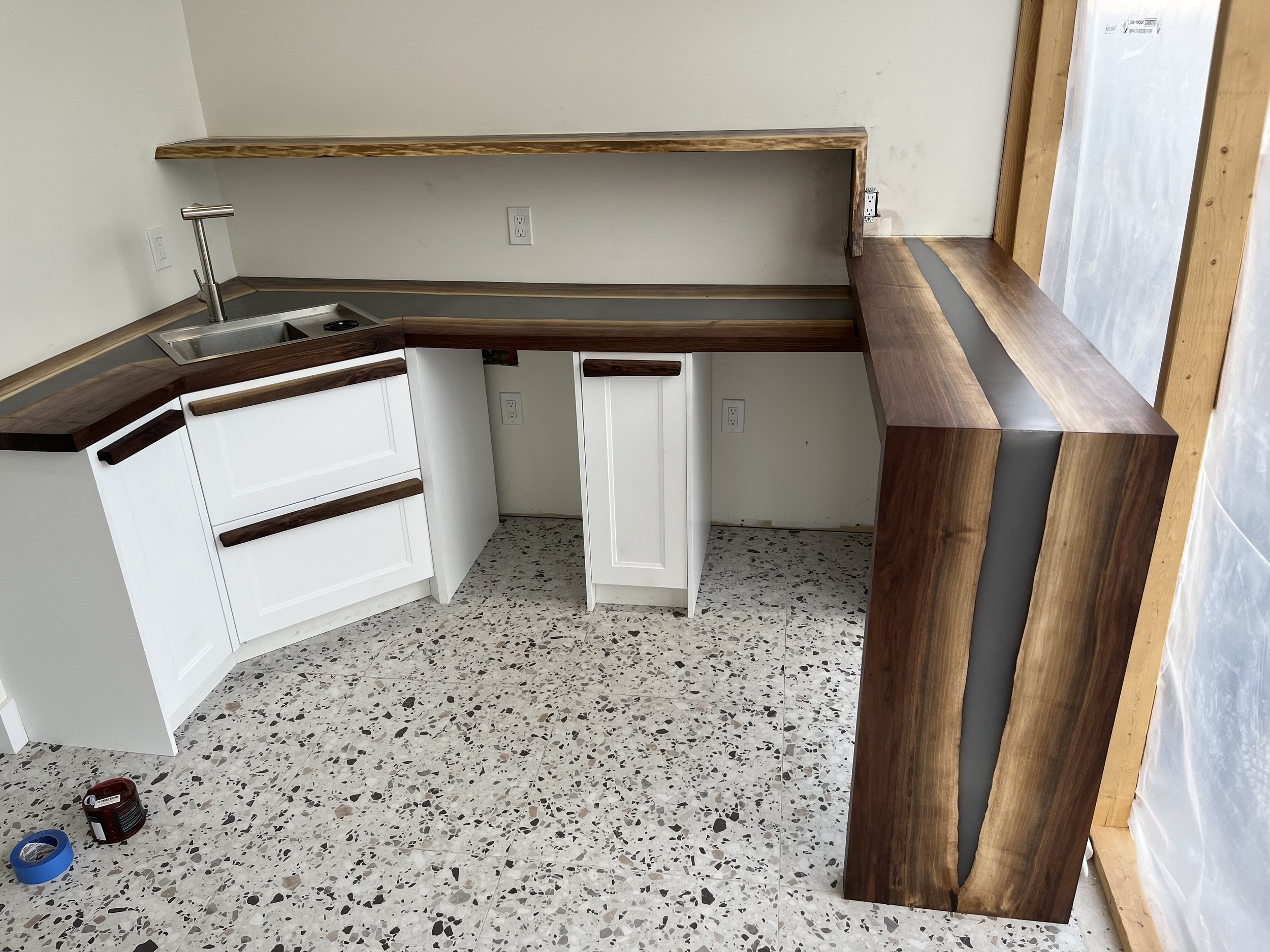 Small modern kitchenette with white cabinets, dark wood countertops, and a stainless steel sink. The space features a terrazzo floor and a matching wood backsplash shelf.