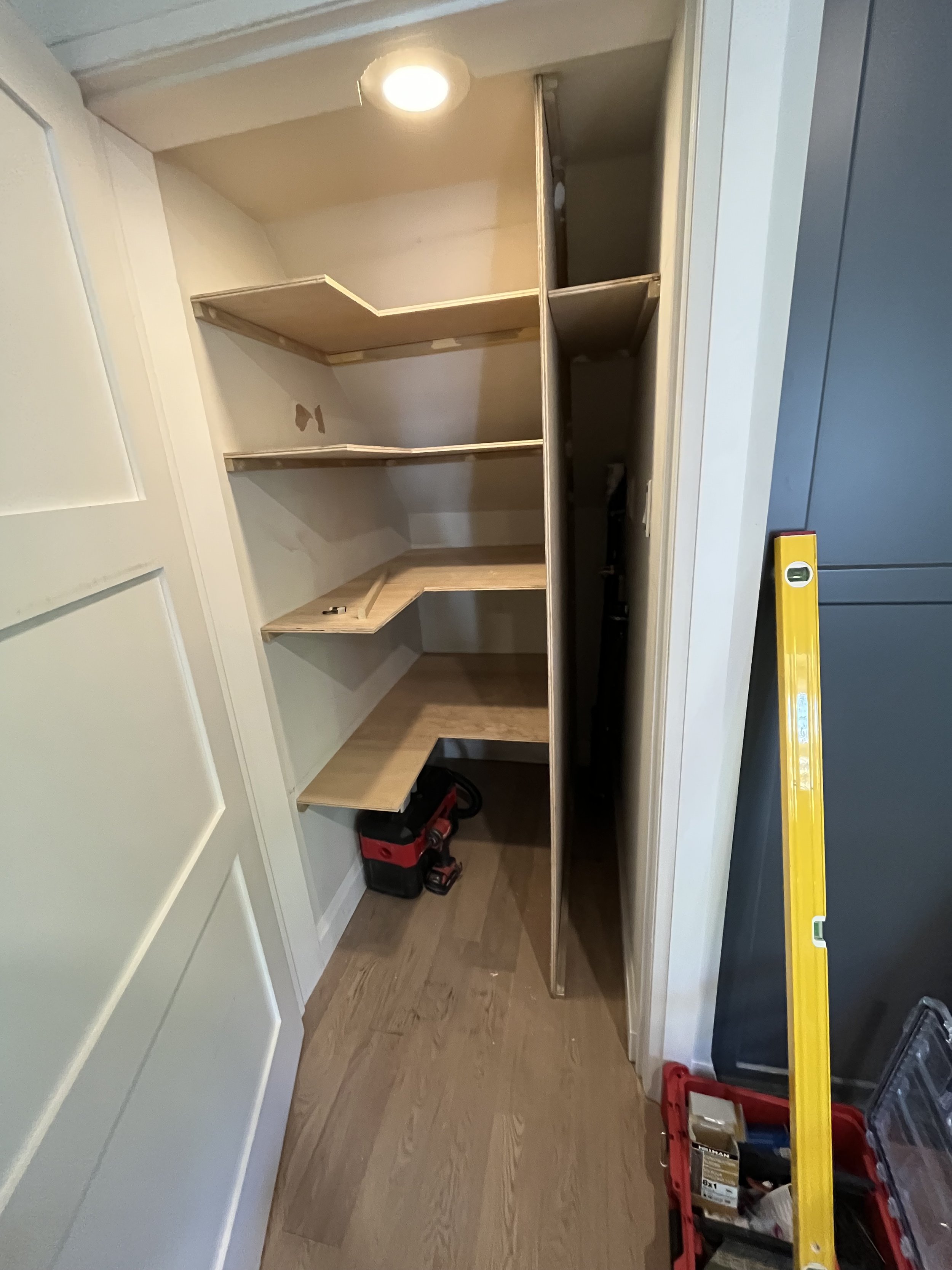 A small closet with wooden shelves installed, a bright overhead light, and some storage items like toolboxes and a level on the floor.