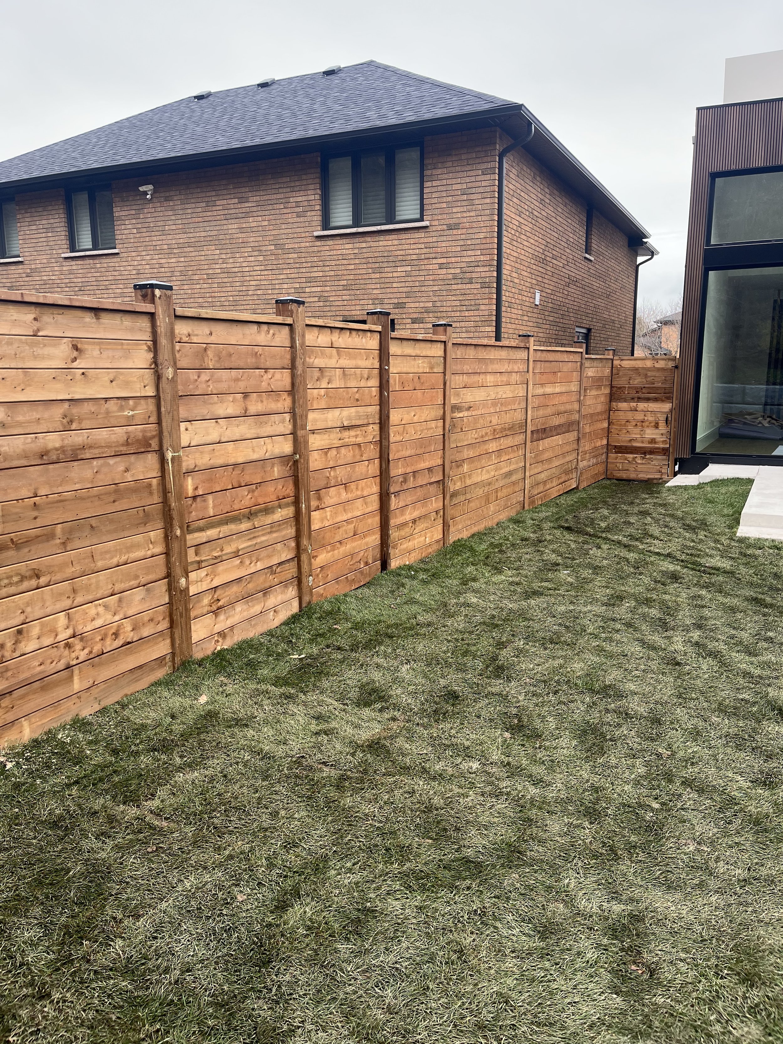 Backyard with wooden fence and grass lawn next to a brick house and modern building, under an overcast sky.