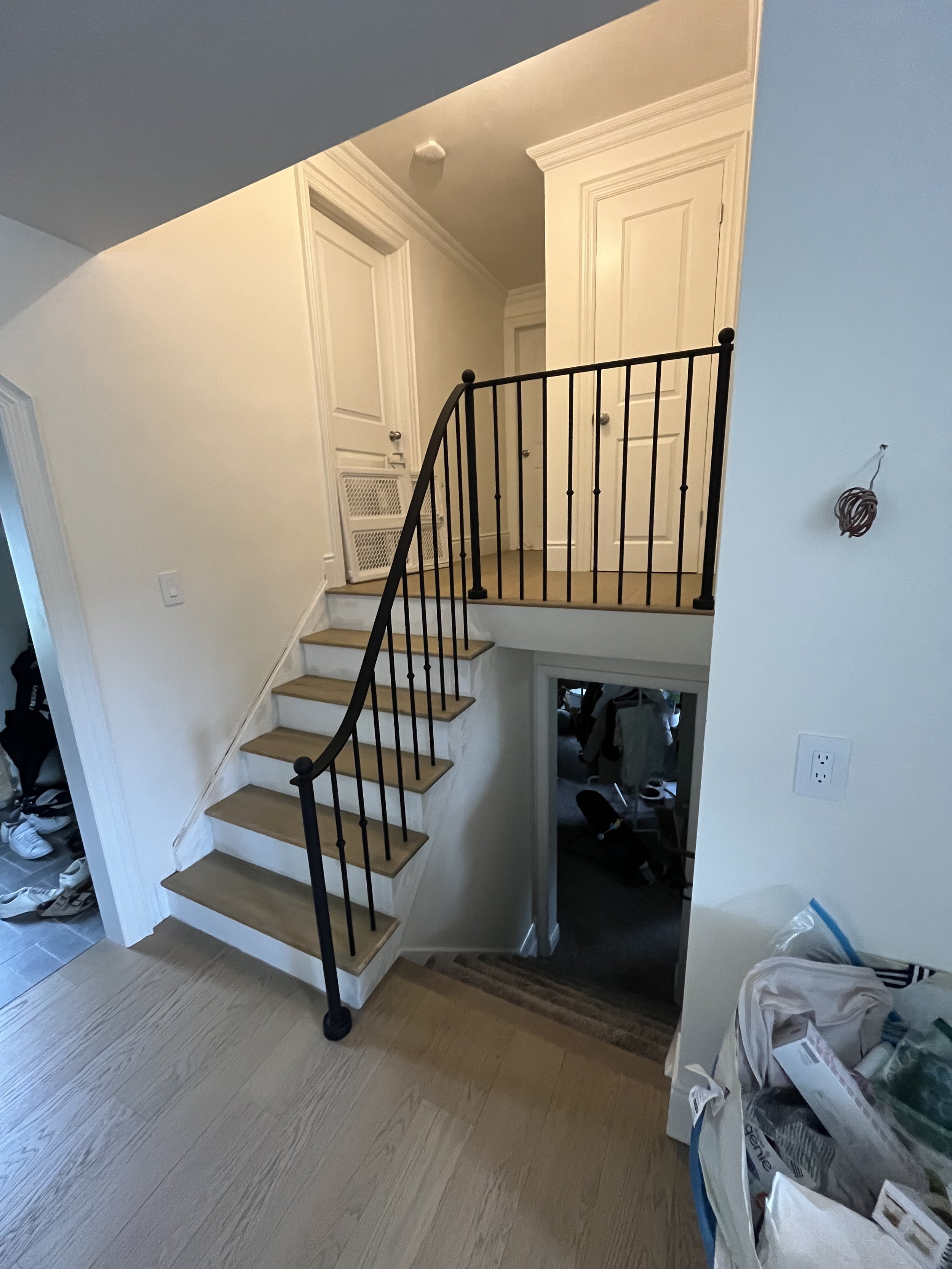 Interior of a house featuring a split staircase with wooden steps and a black metal railing. The stairs lead up to a landing with two white doors and down to a lower level room filled with clothes. A large laundry bag and a wall light switch are visi