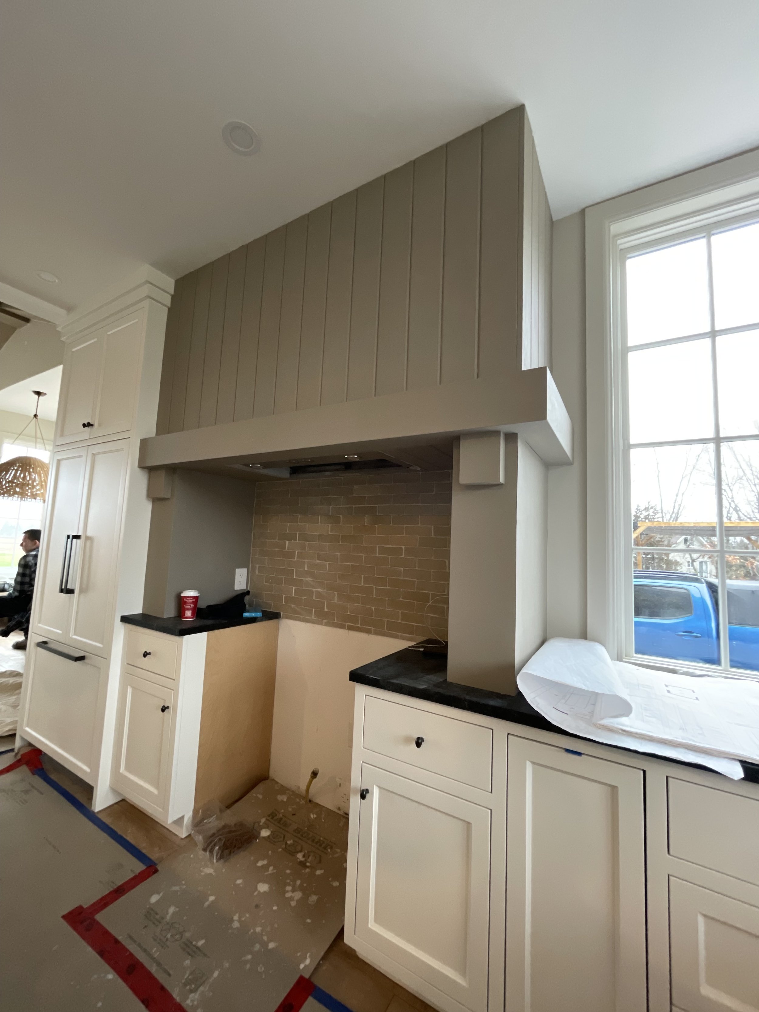 Kitchen under construction with beige cabinetry, a hood range, and a brick-style backsplash. There are some construction materials on the floor and a large window on the right side.
