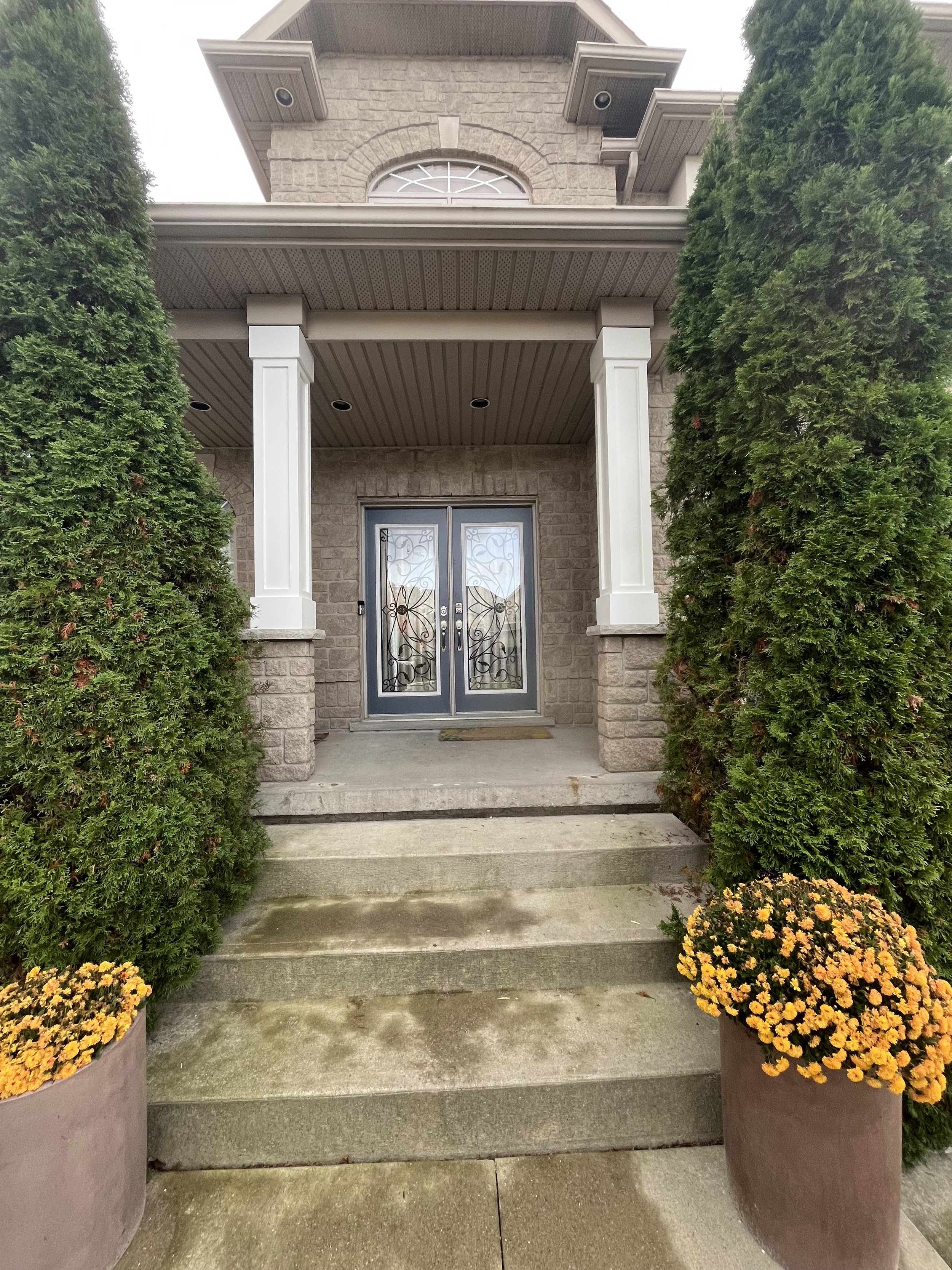 Front entrance of a house with double doors, stone facade, white columns, large shrubs, and potted yellow flowers on concrete steps.