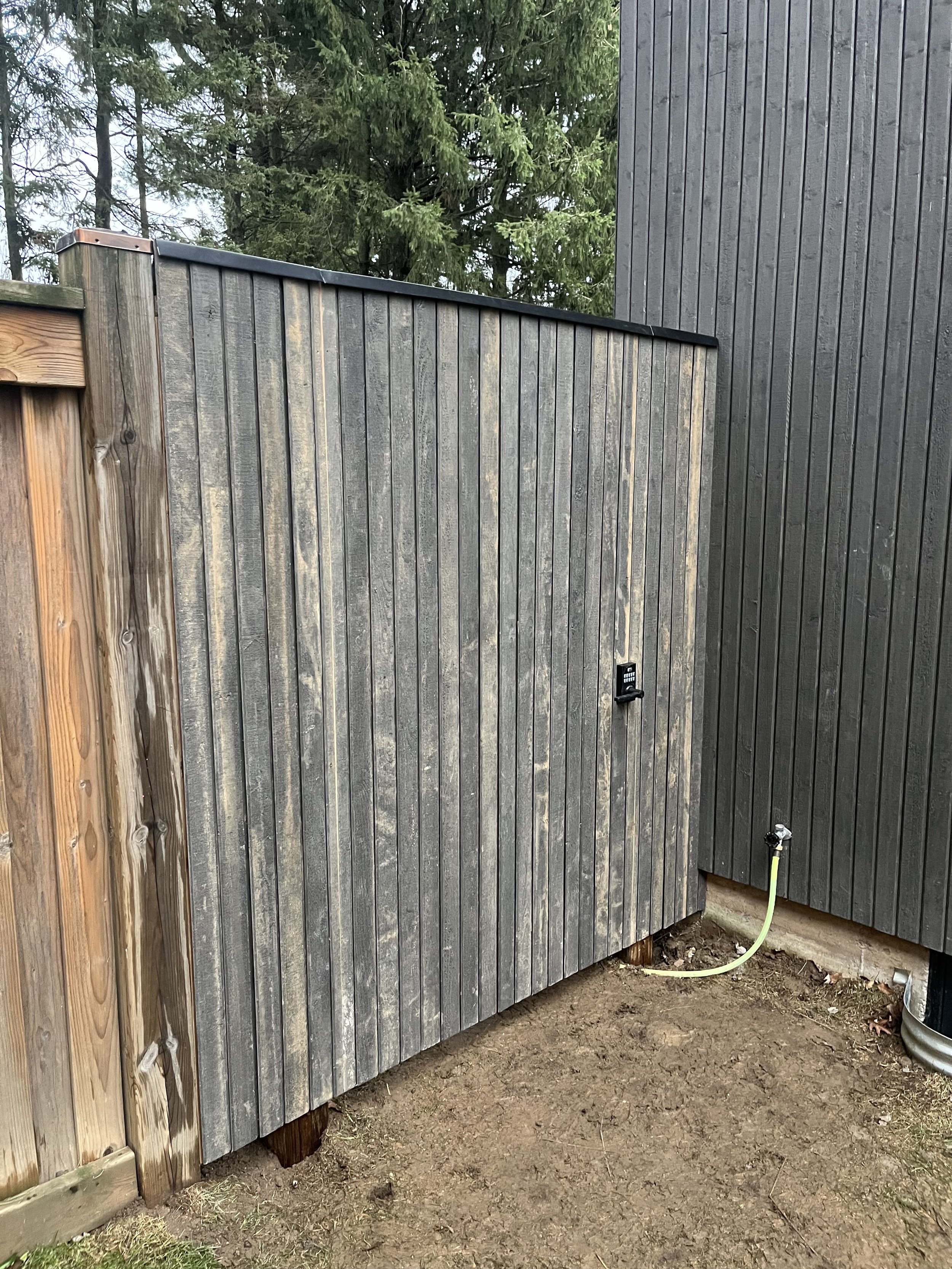 Wooden fence with a gate, adjacent to a gray building, and a hose attached to a tap on the wall.