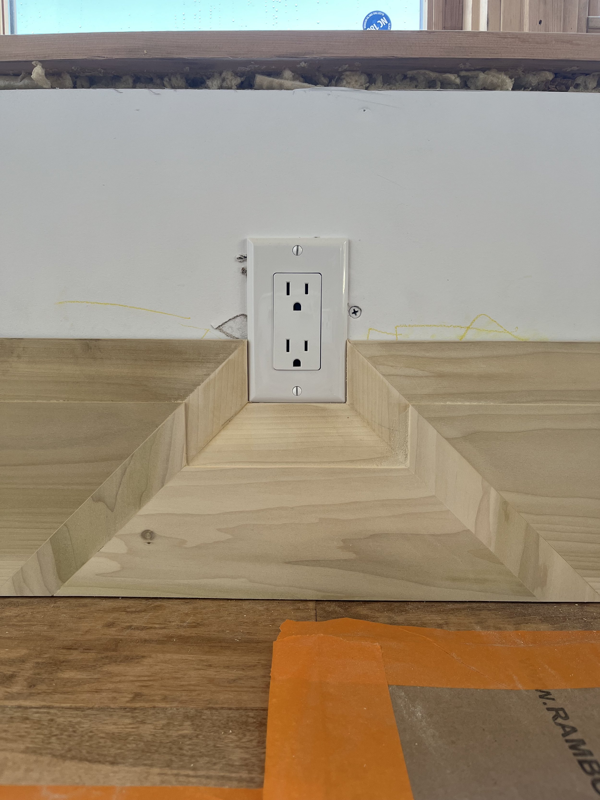 White electrical outlet installed on a wall with unfinished wooden paneling and exposed insulation above it. The wooden paneling is arranged with mitered corners. The edge of a wood surface with orange tape is visible at the bottom.
