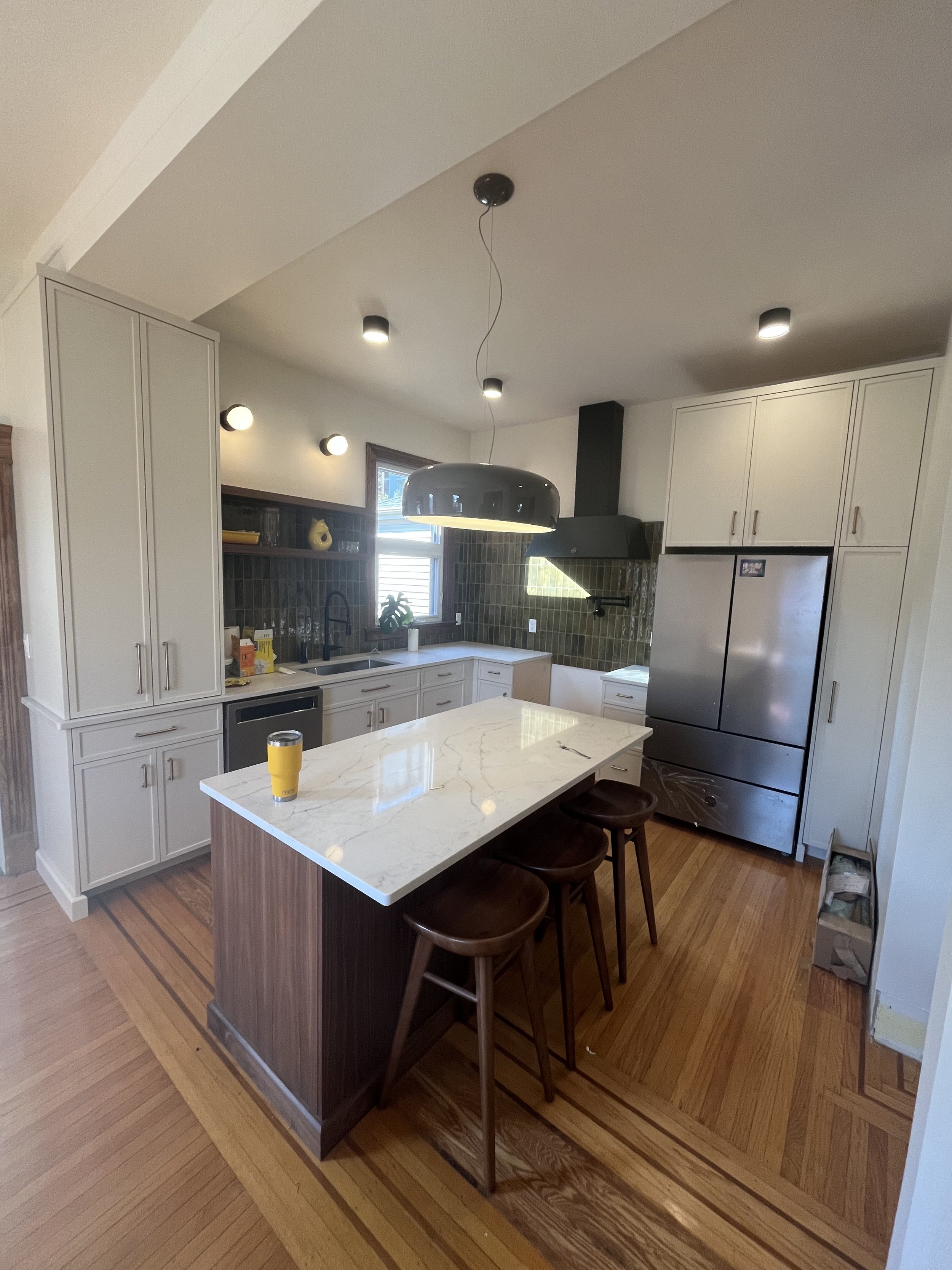 Modern kitchen with white cabinets, stainless steel appliances, and a large central island with a marble countertop. Three wooden stools are placed at the island. The floor is wooden, and a pendant light hangs from the ceiling. A glass with an orange