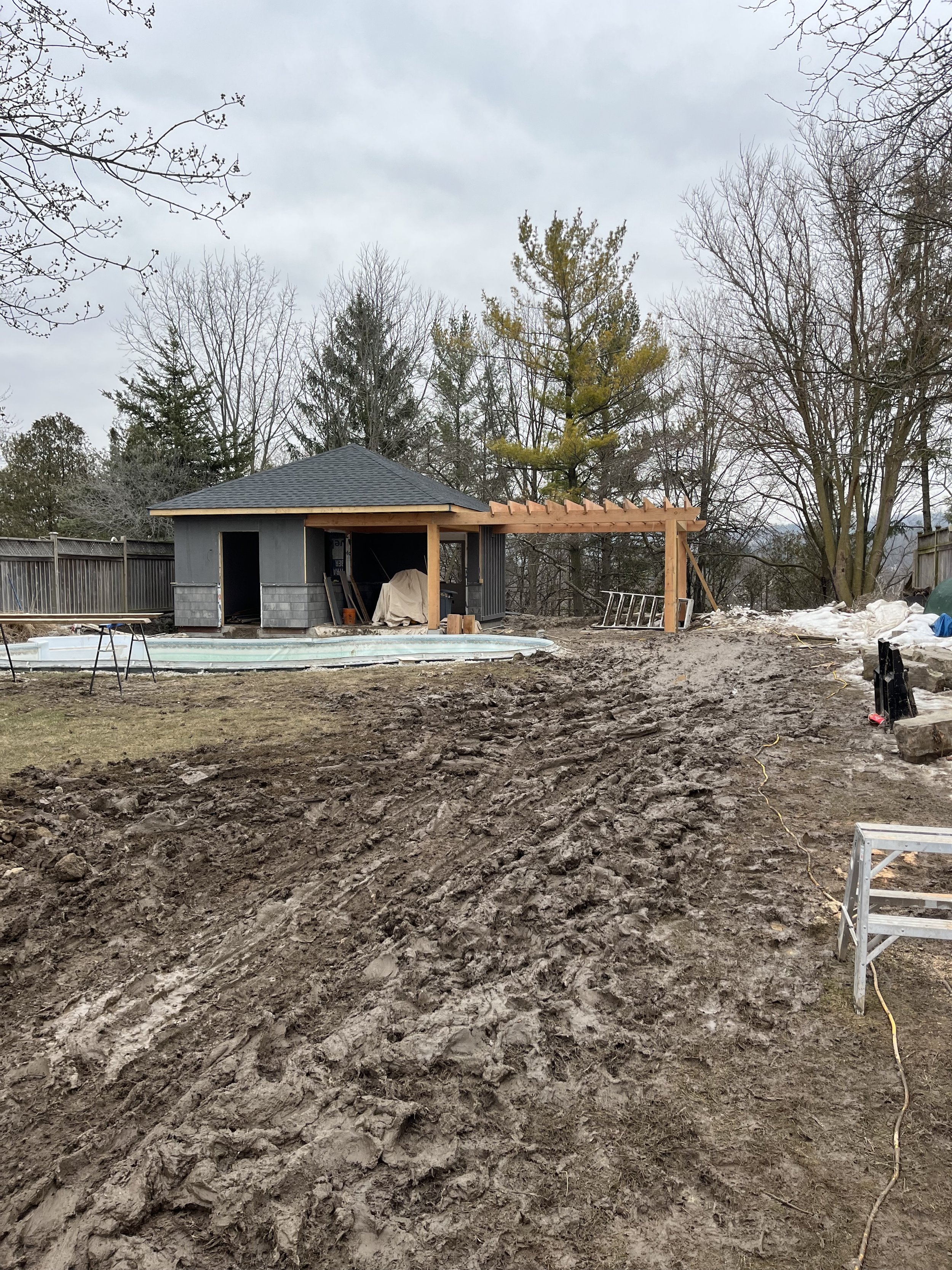 Outdoor construction site with a small building under construction. Features a partially built pergola, a covered swimming pool on the left, muddy ground, some tools, and trees in the background.