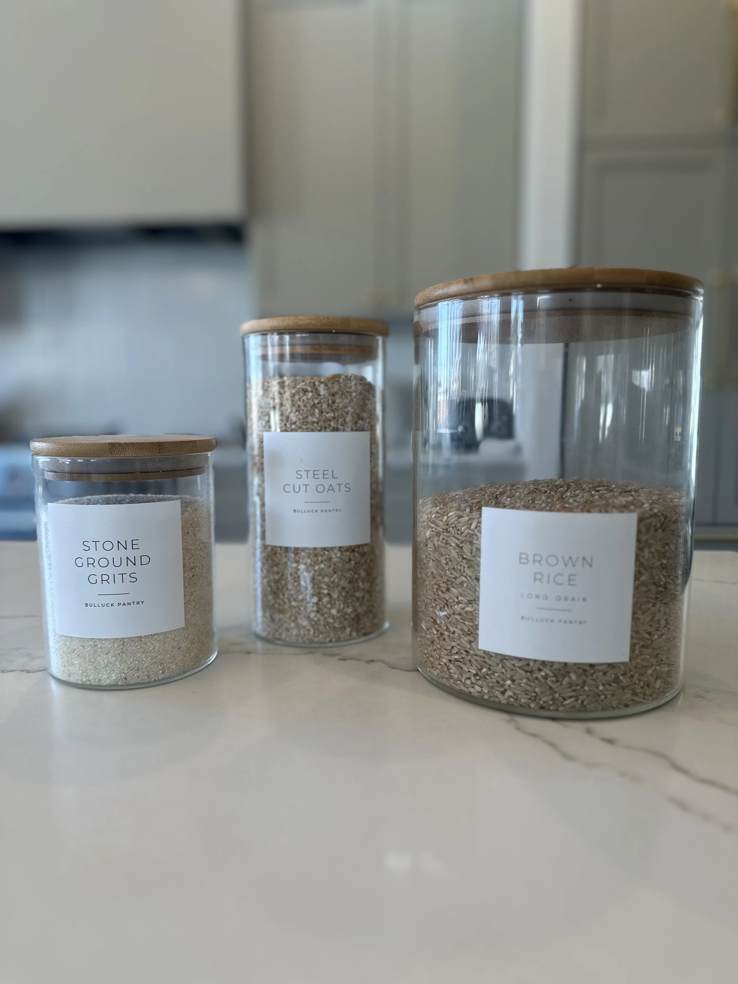 Three glass jars with wooden lids on a countertop, labeled as "Stone Ground Grits," "Steel Cut Oats," and "Brown Rice."