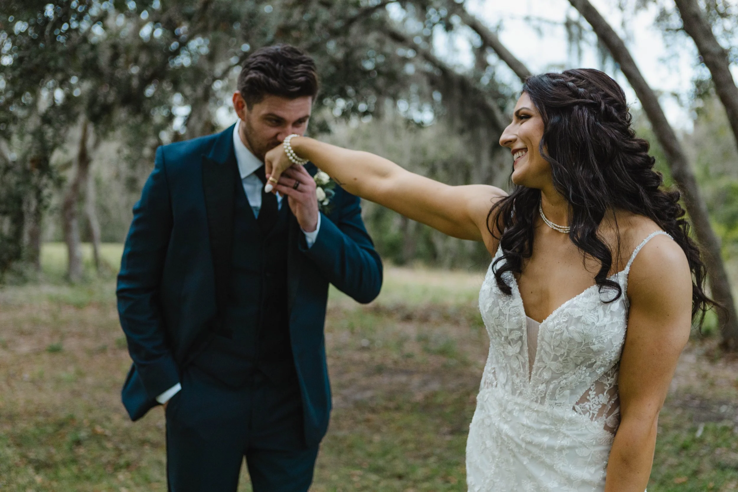 A man in a suit kisses a woman in a lace wedding dress on the cheek, with a woman smiling in the background outdoors among trees.