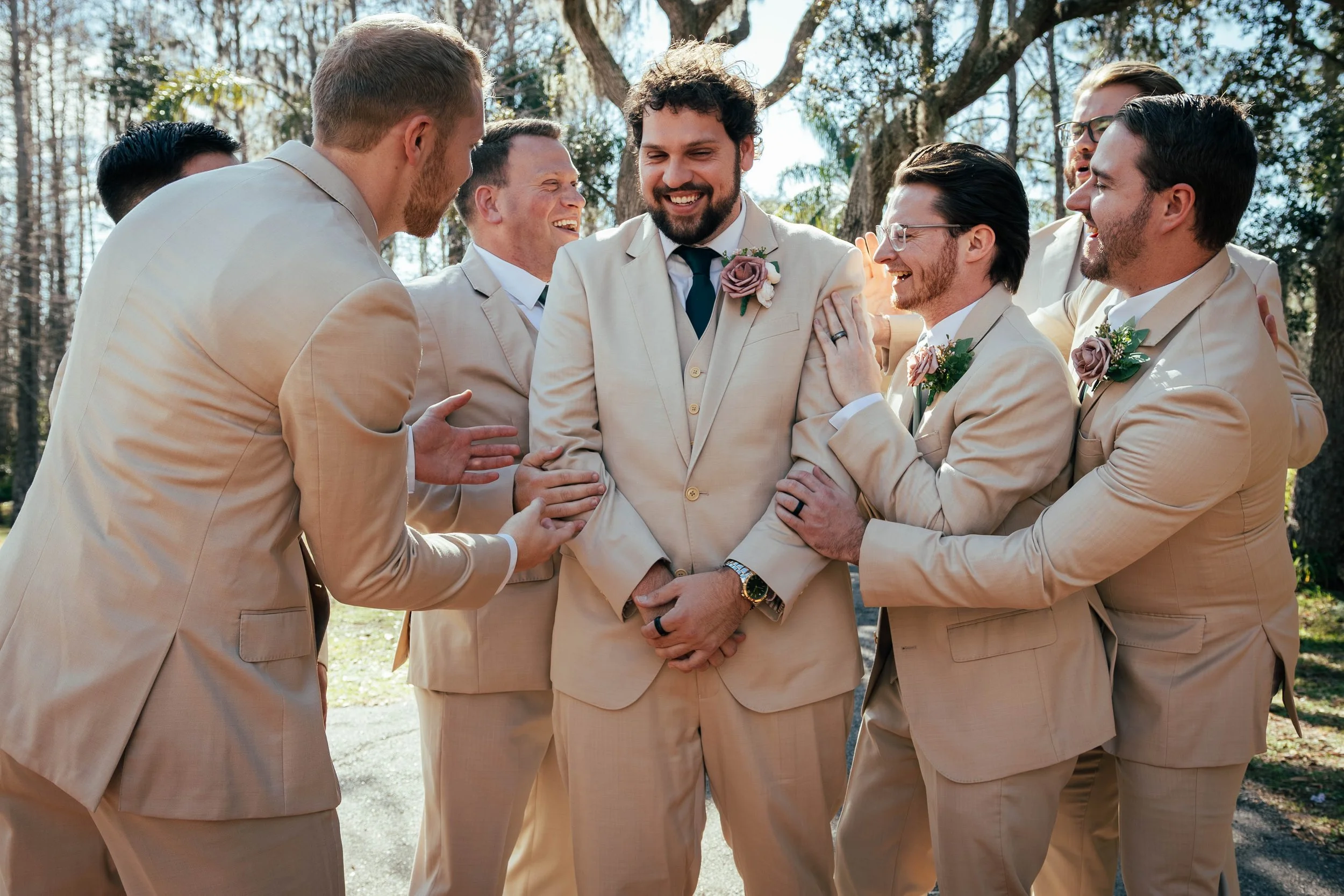 Group of men in beige suits, smiling and celebrating, outdoors with trees in the background, during daytime.