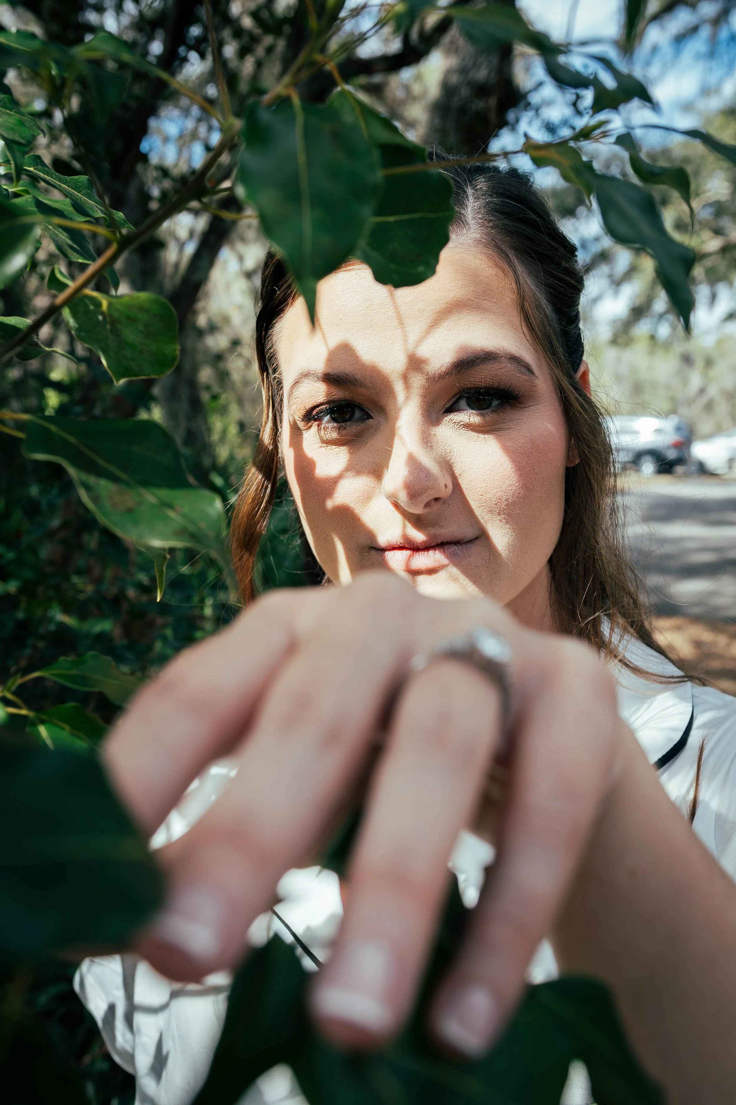 A woman with light skin, dark brown hair, and makeup is outdoors among green foliage, reaching towards the camera with her hand, which is in focus, while her face is slightly blurred in the background. Sunlight creates shadows on her face from the le