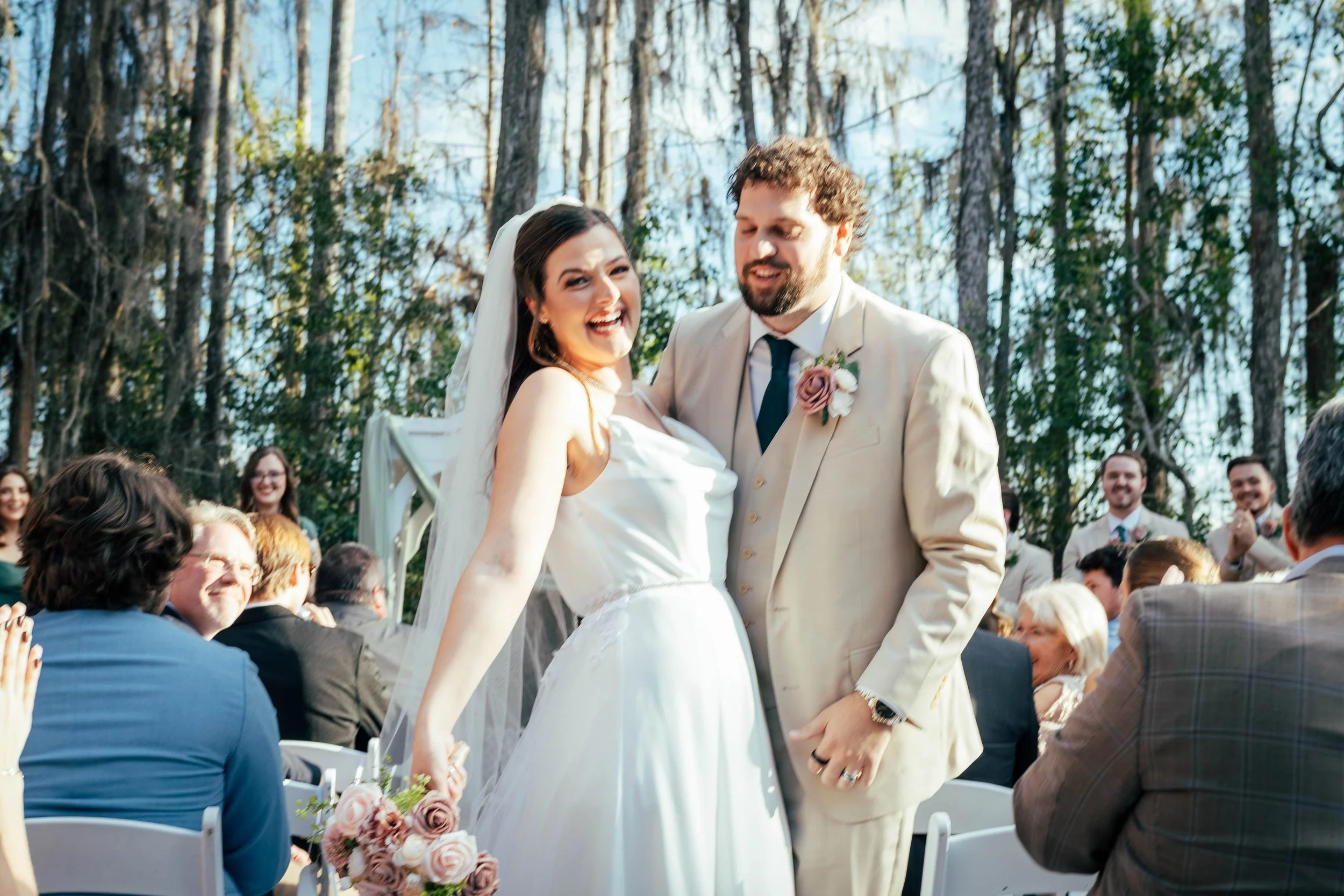 A bride and groom smiling and holding hands during an outdoor wedding ceremony, surrounded by seated guests, with trees in the background.