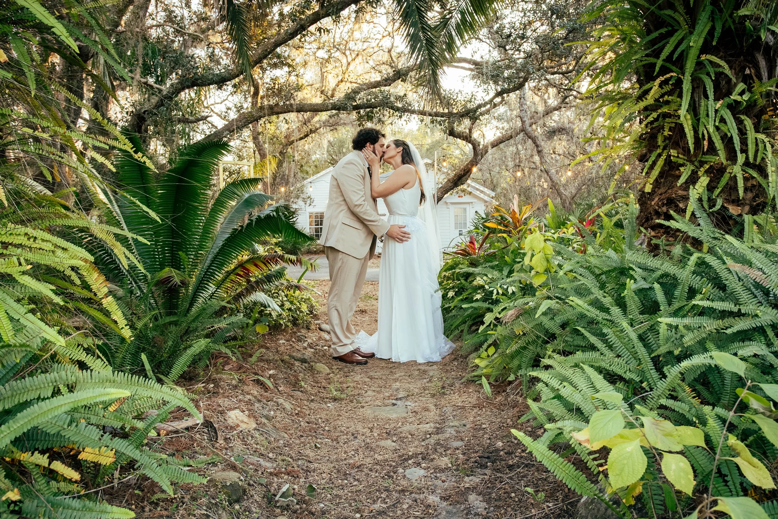 A couple shares a kiss in a garden setting surrounded by lush green tropical plants and trees, with sunlight filtering through the branches.