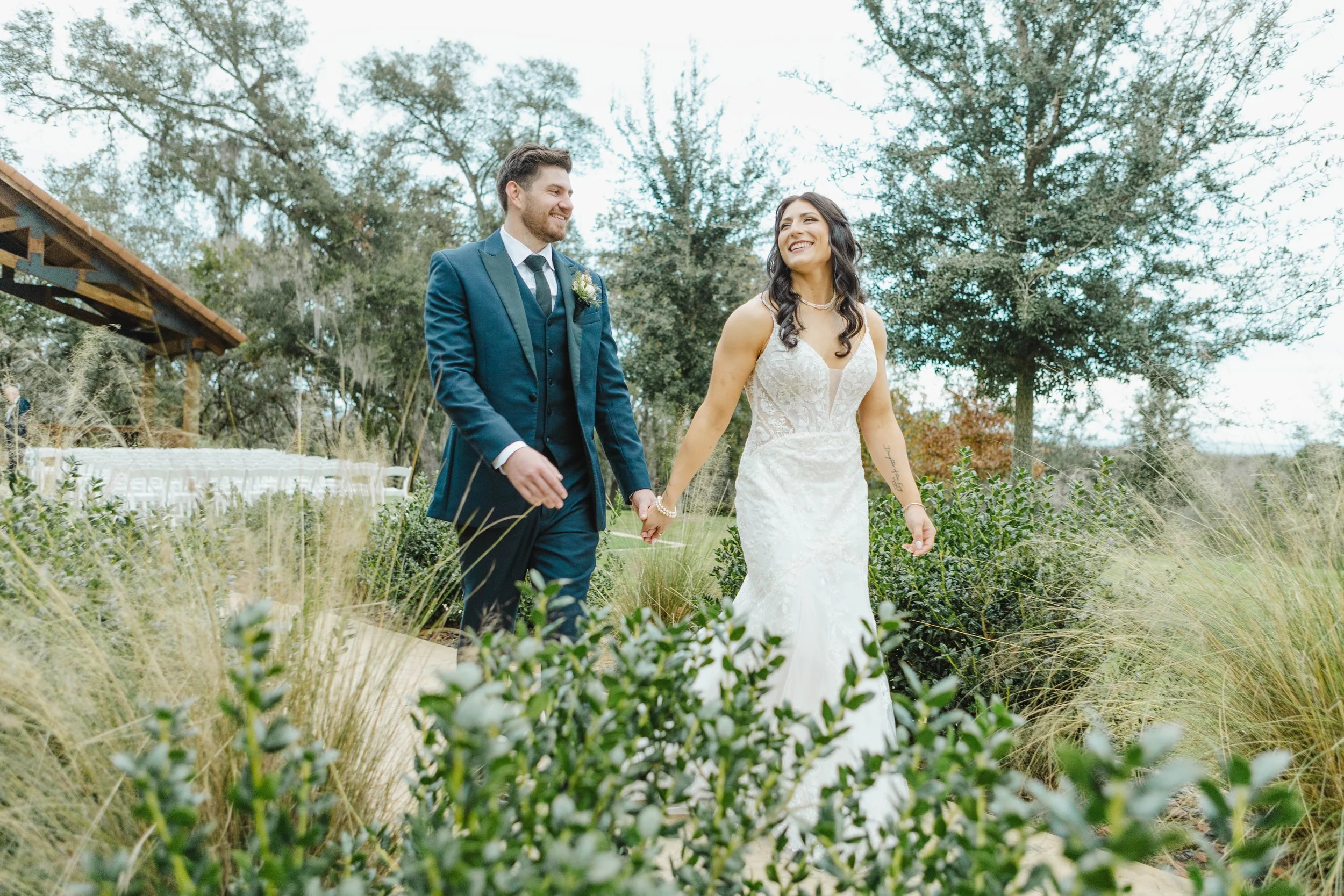 A bride and groom walking hand in hand outdoors during a wedding ceremony surrounded by greenery and trees.